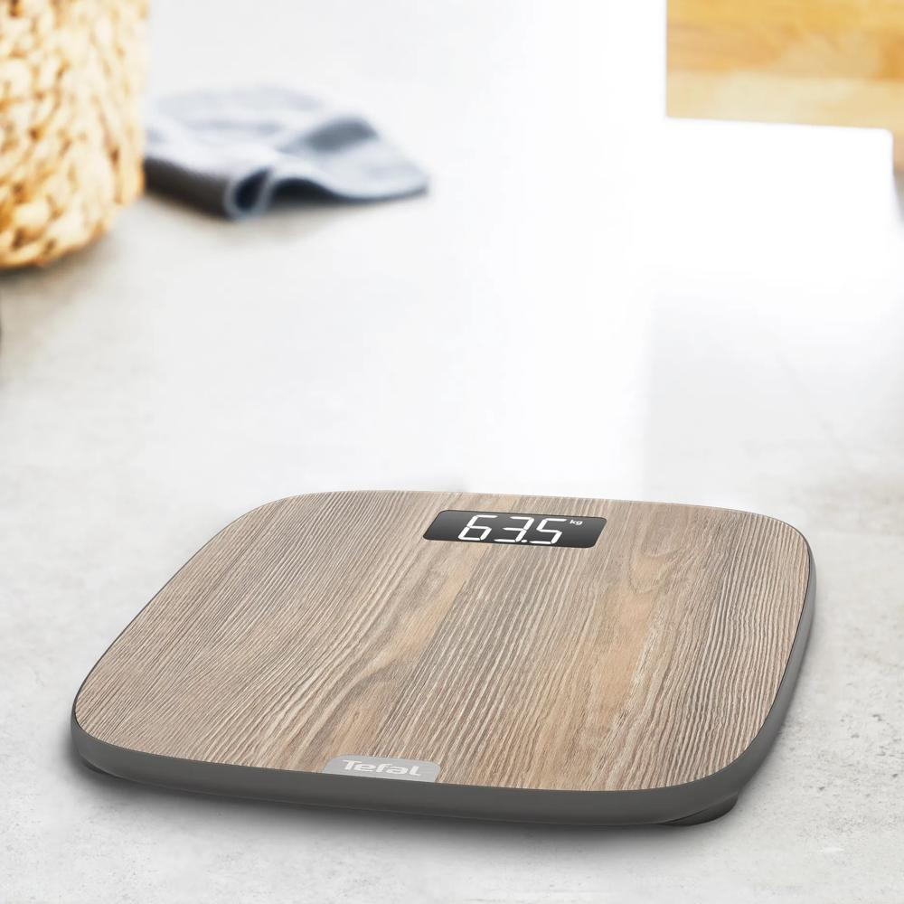 Bilancia pesapersone Tefal con motivo in legno su pavimento chiaro, segna 63,5 kg.