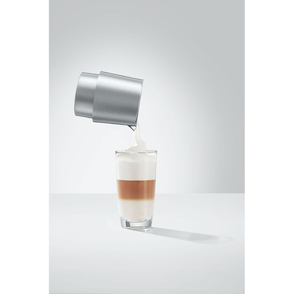 Il latte viene versato da una lattiera d'argento in un bicchiere di latte macchiato su un tavolo bianco.