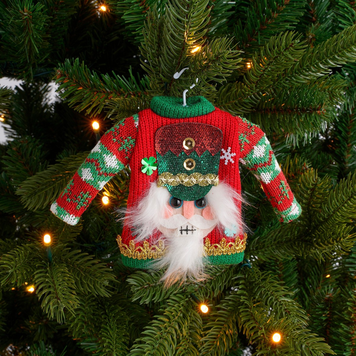 A nutcracker Christmas sweater ornament hangs on a decorated Christmas tree.