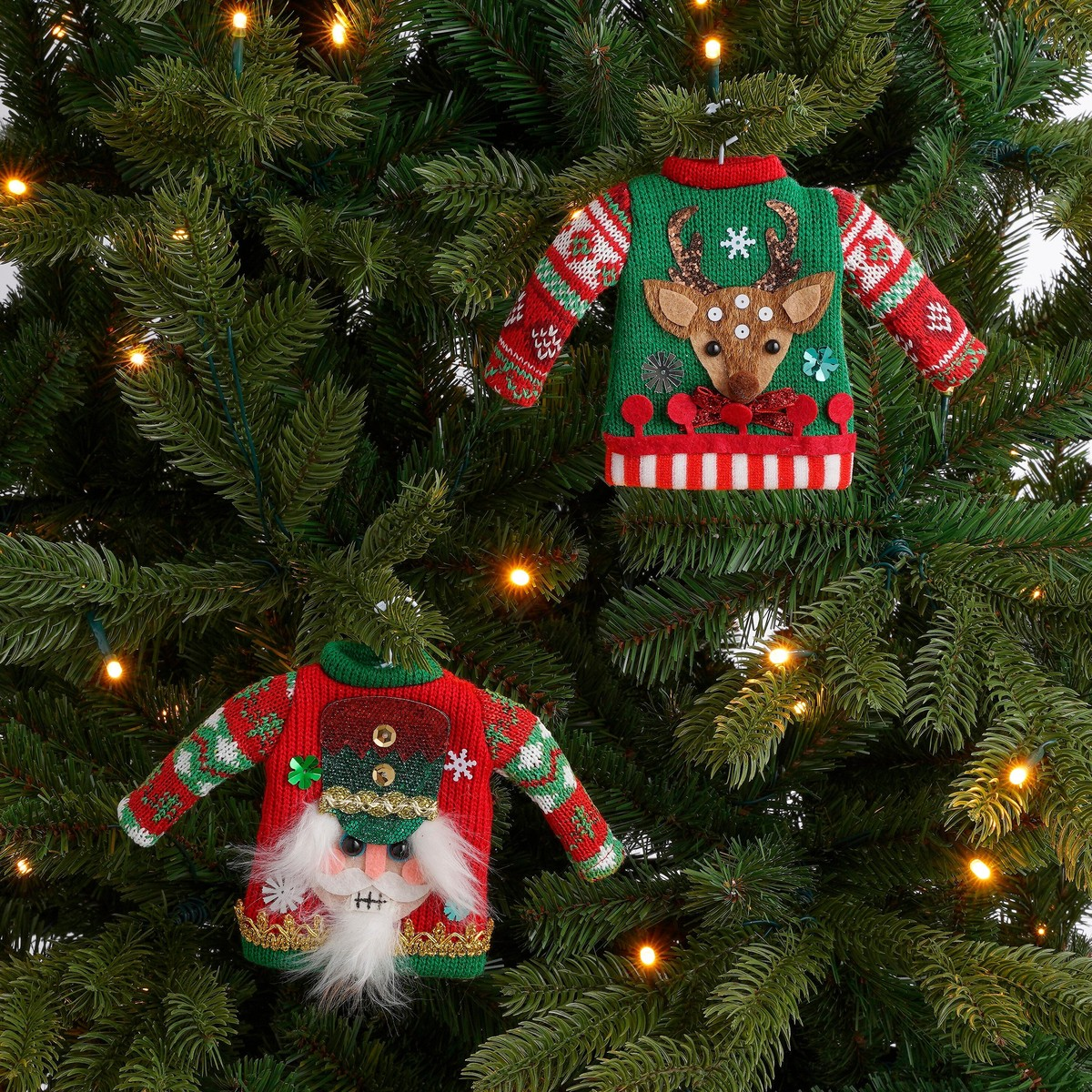 Two small knitted holiday sweater ornaments, one with a reindeer and one with a nutcracker, hang in a tree.