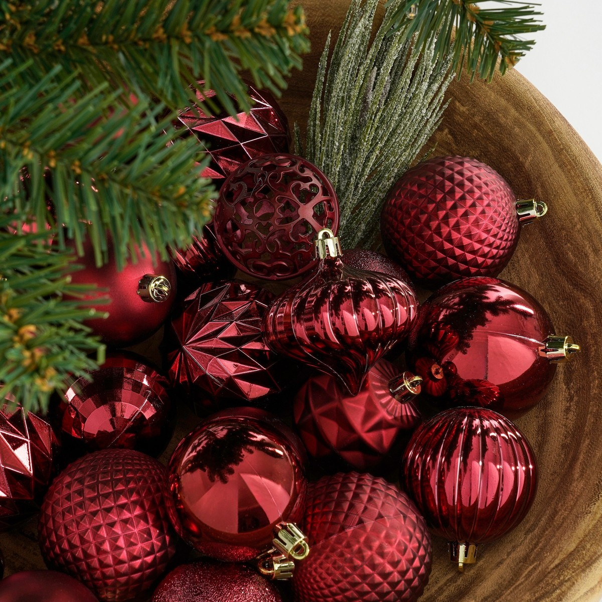A wooden bowl filled with red Christmas ornaments and pine branches.