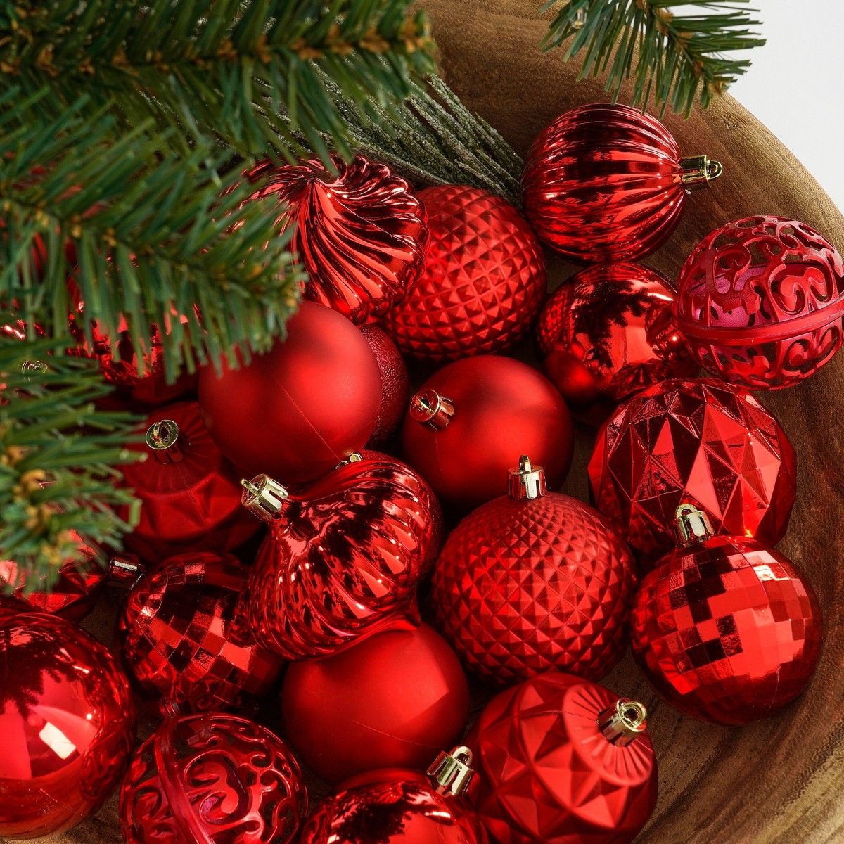 A wooden bowl filled with shiny red Christmas ornaments and pine branches.