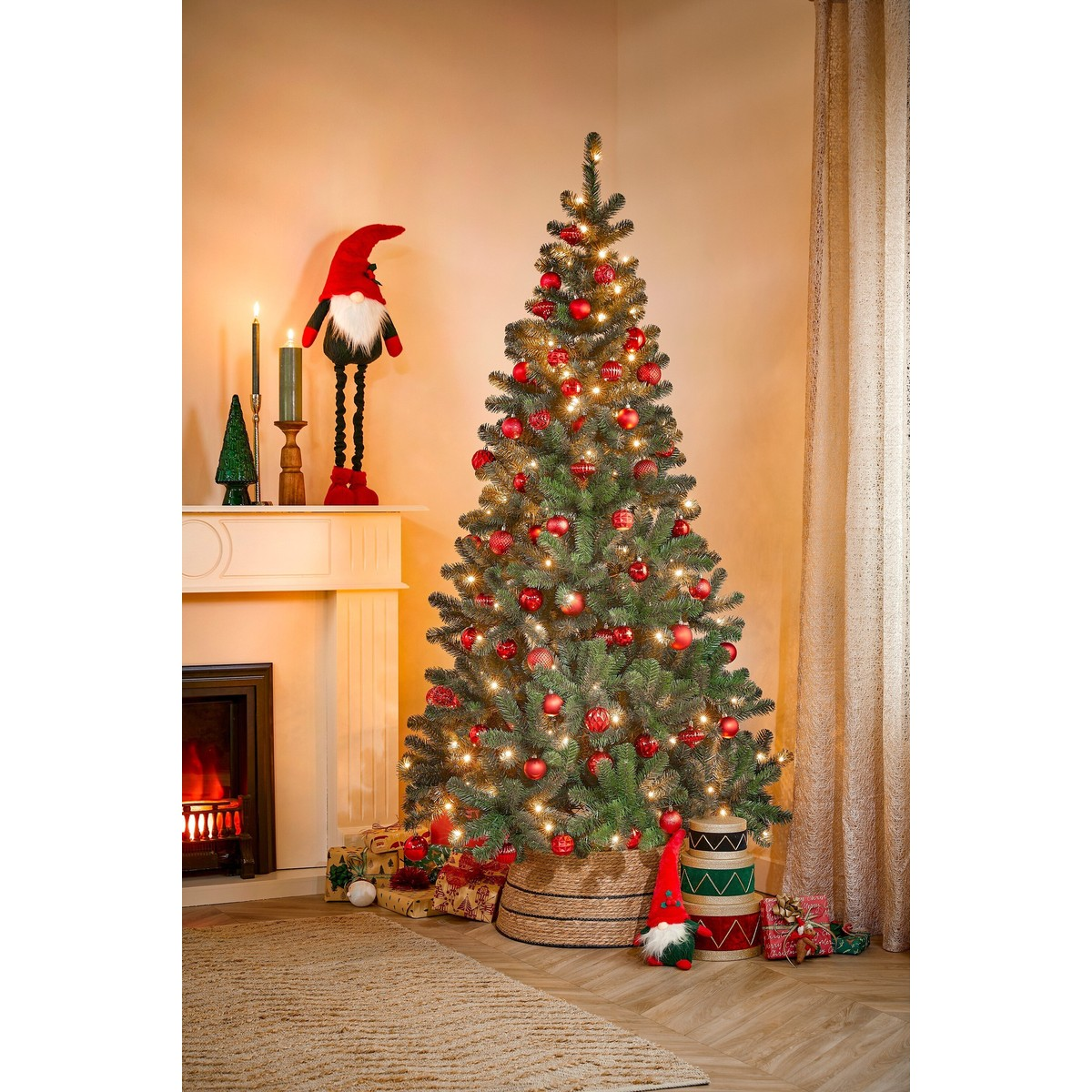 An illuminated Christmas tree decorated with red baubles, gifts, and a fireplace in the background.