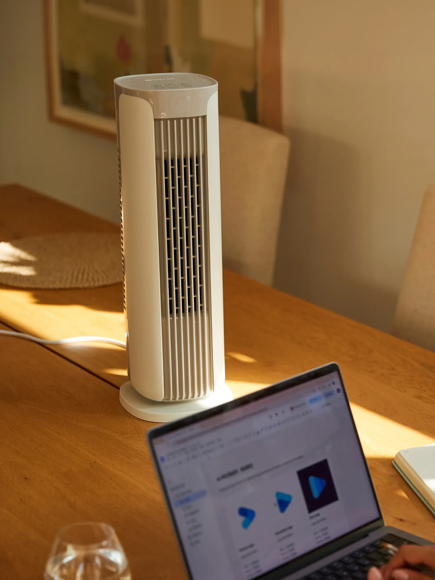 Ein weißer Turmventilator steht auf einem Holztisch neben einem Laptop in einem sonnigen Raum.
