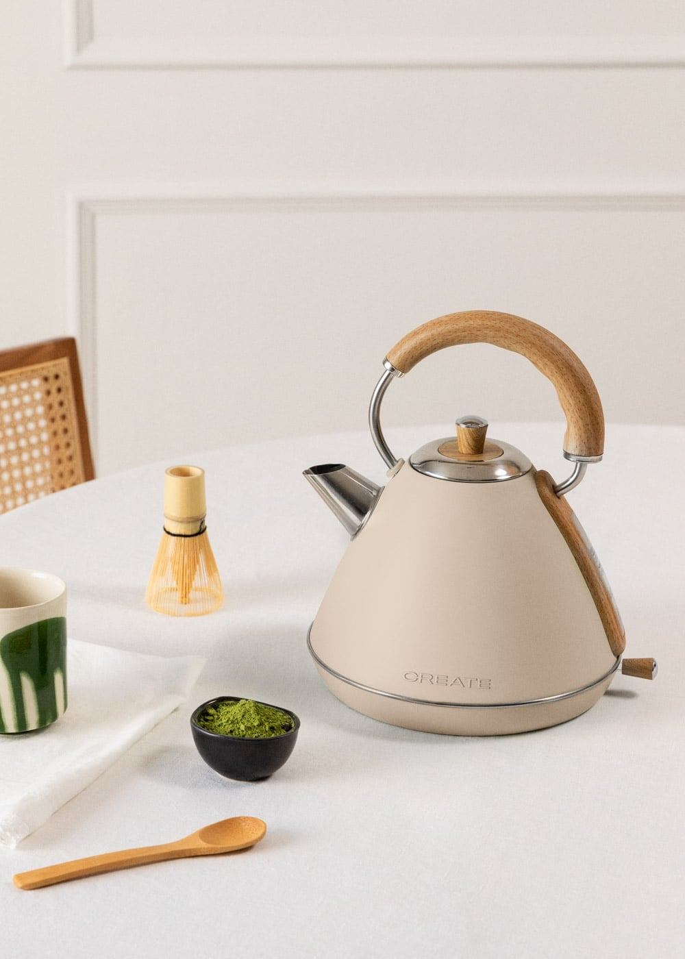 Un hervidor beige, bol de matcha, batidor de madera y cuchara sobre un mantel blanco.