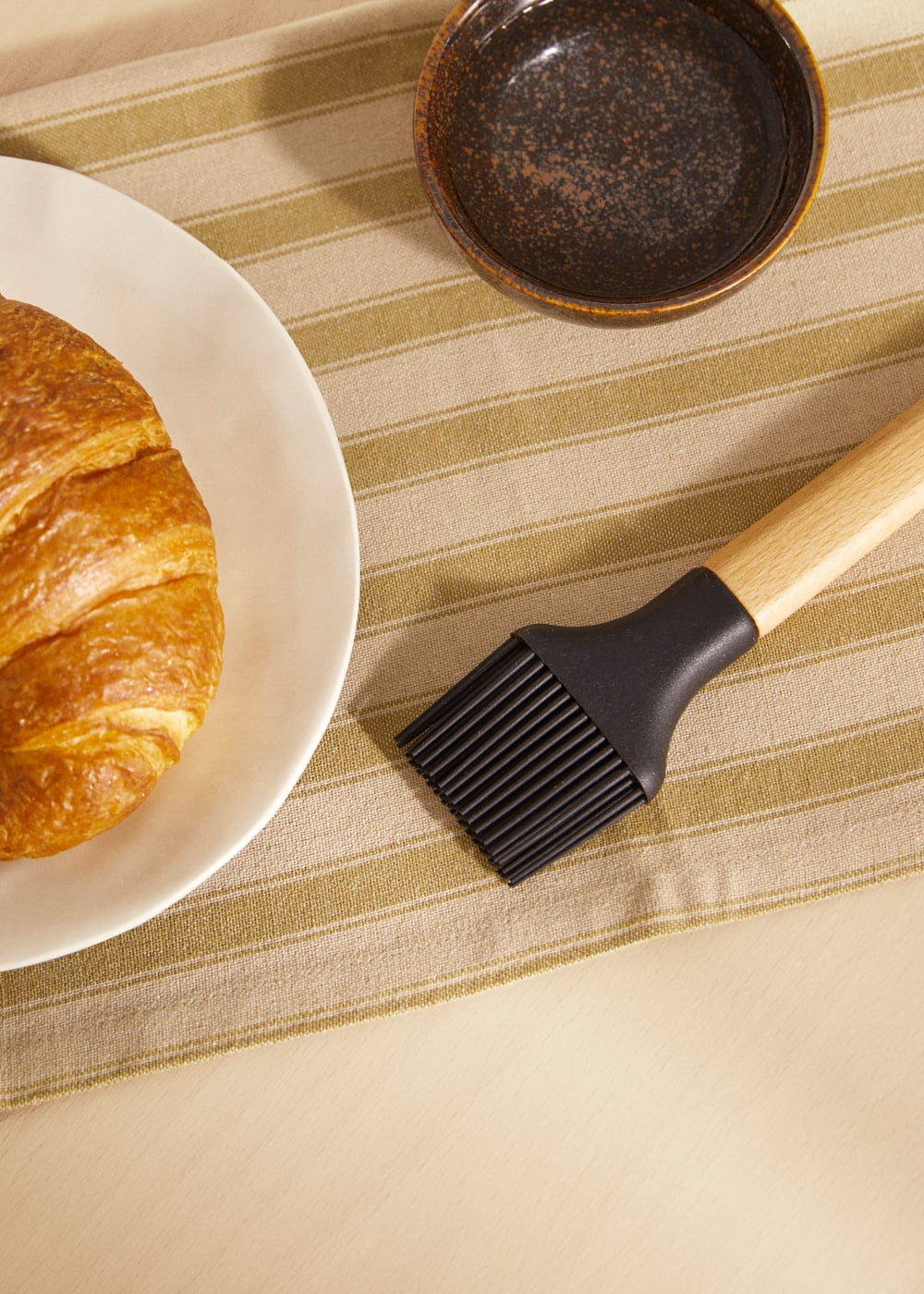 Un cruasán en un plato blanco, una brocha de repostería con mango de madera y un cuenco oscuro en un mantel a rayas.