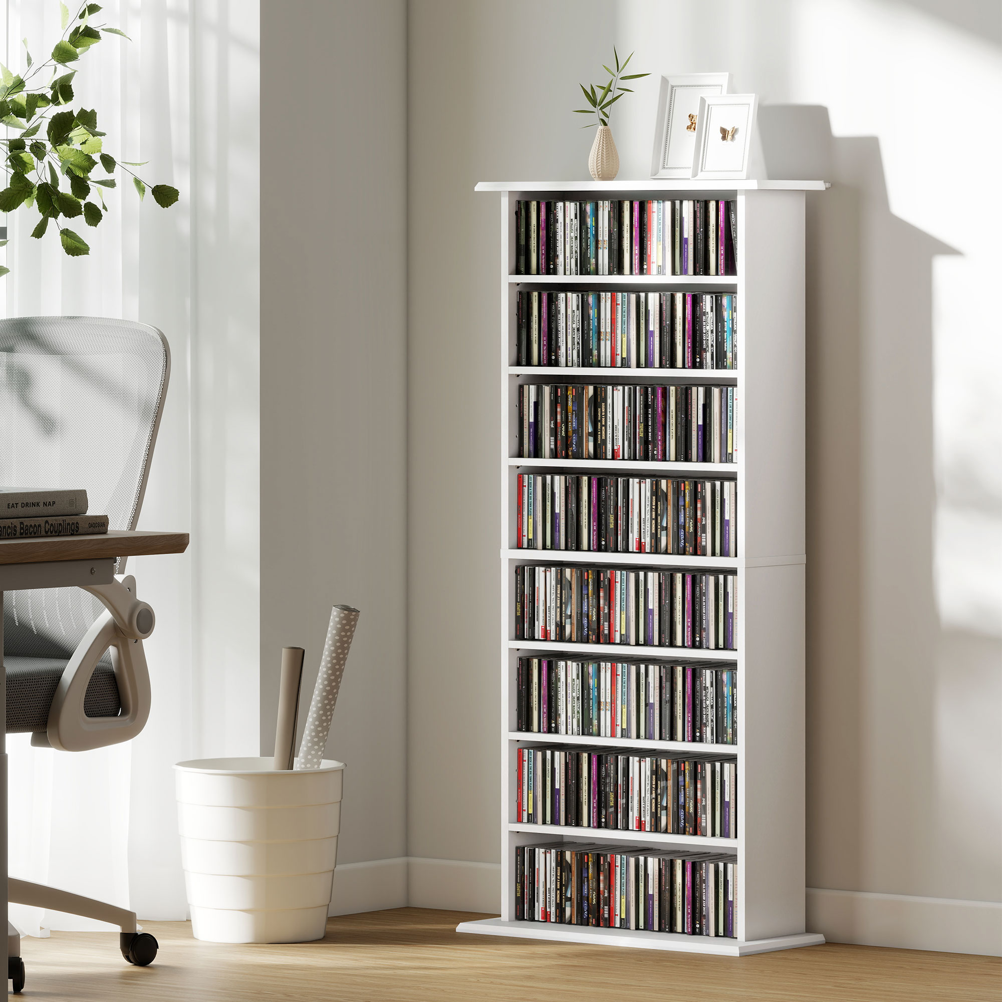 Una estantería blanca para CD llena de CD en una habitación luminosa junto a un escritorio y papelera.