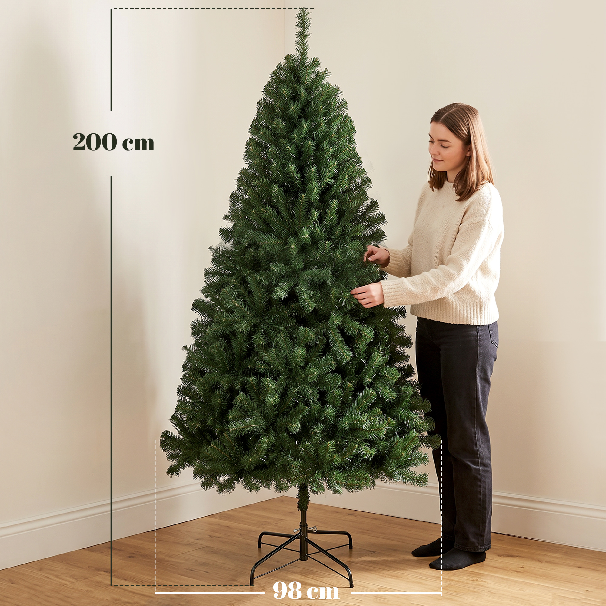 Una mujer con un jersey claro se encuentra junto a un árbol de Navidad artificial de 200 cm.
