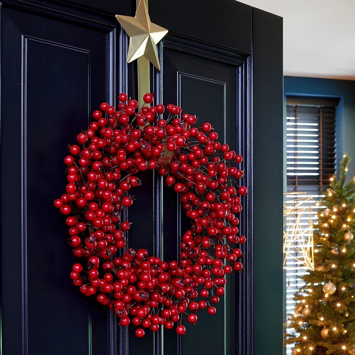 A red berry wreath hangs on a black door beneath a golden star, with a Christmas tree in the background.