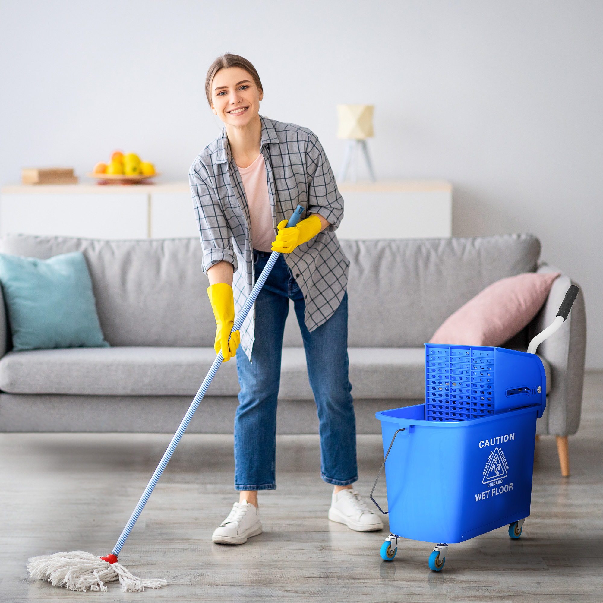 Una mujer con guantes amarillos friega el suelo de una sala con una fregona azul junto a un cubo.