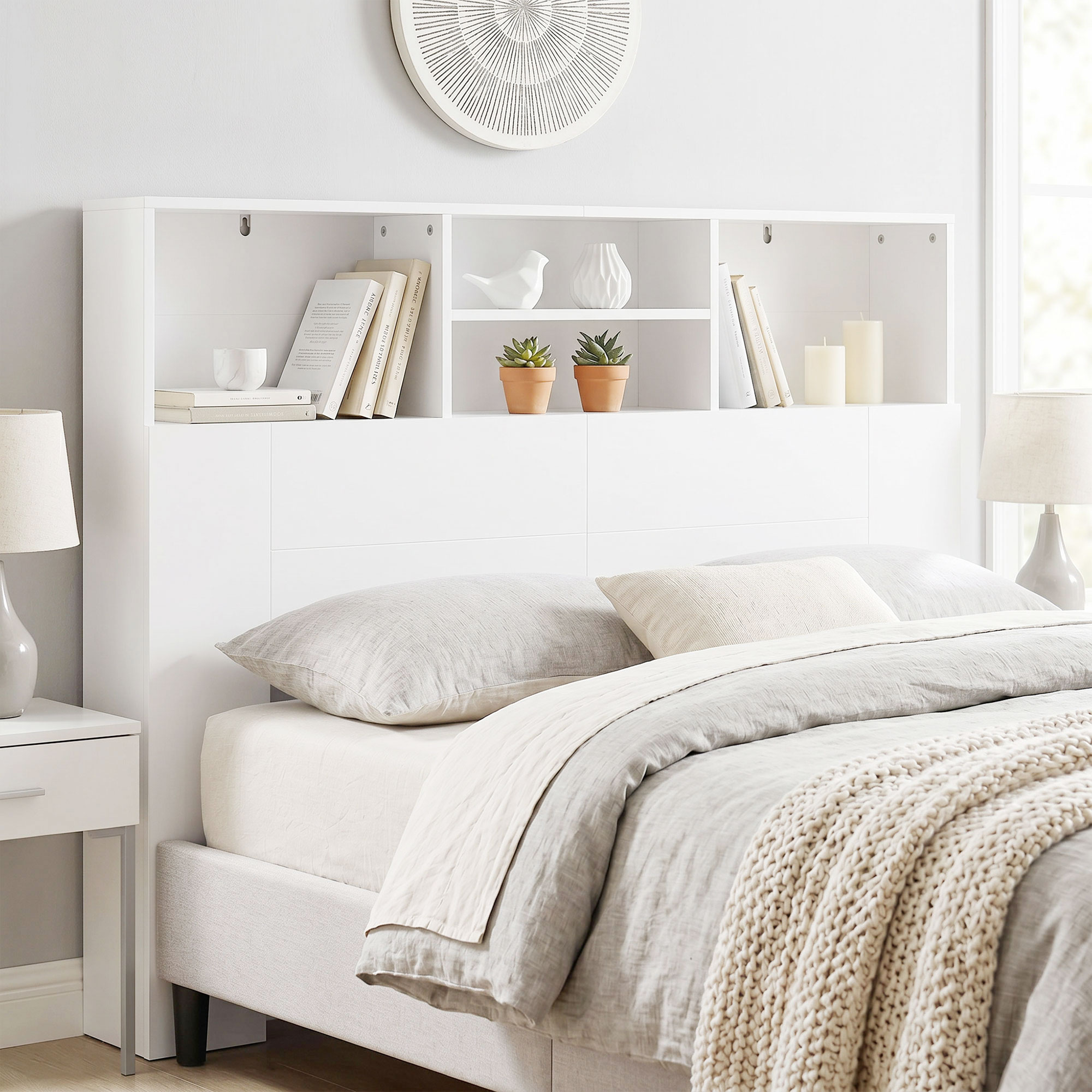 Cama blanca con cabecera de estantería integrada con libros, plantas y decoración, flanqueada por mesitas.