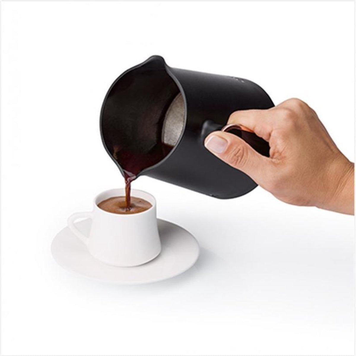 Eine Hand gießt Kaffee aus einer schwarzen Kanne in eine weiße Tasse auf einem weißen Unterteller.