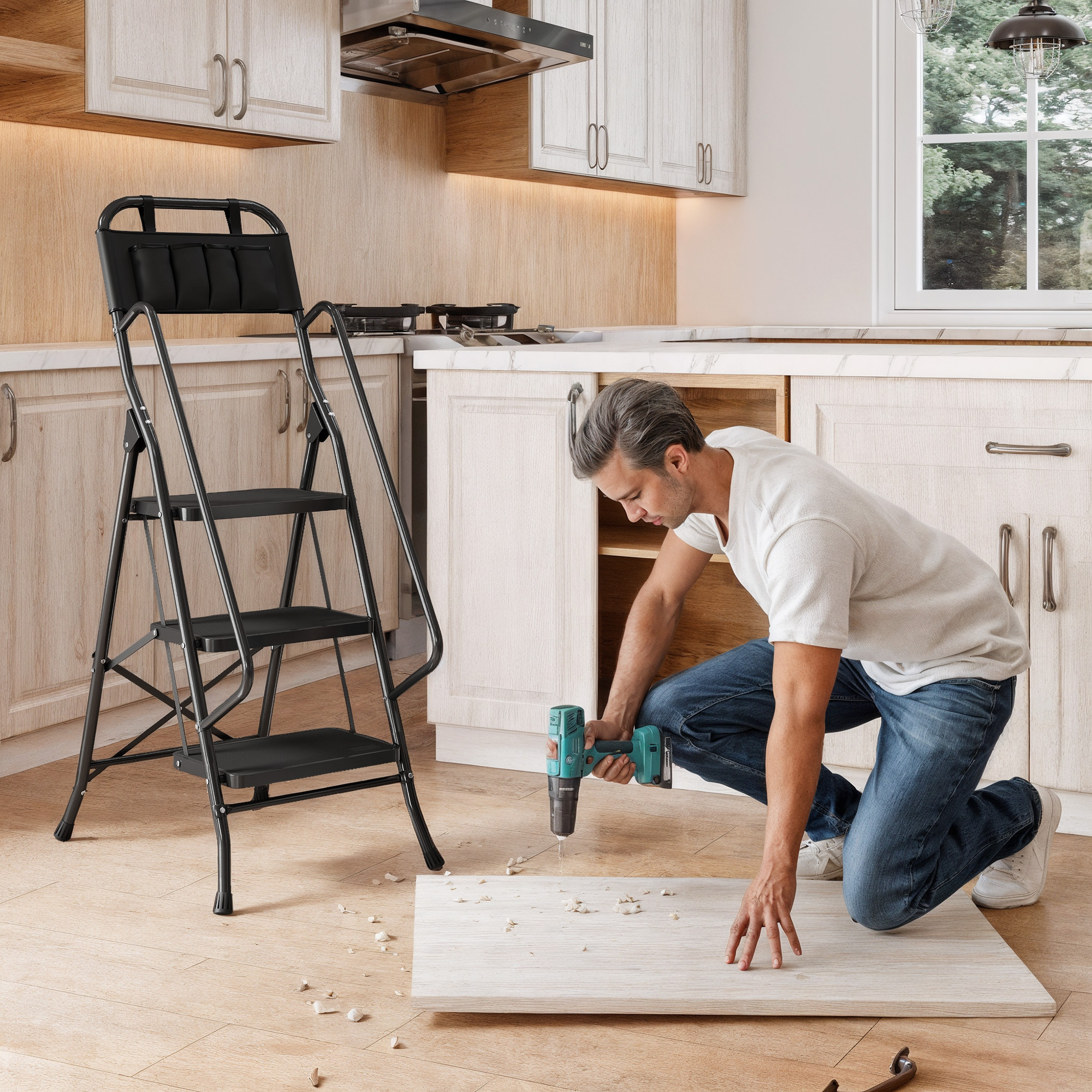 Un hombre se arrodilla en el suelo de una cocina taladrando una tabla de madera, con una escalera a la izquierda.