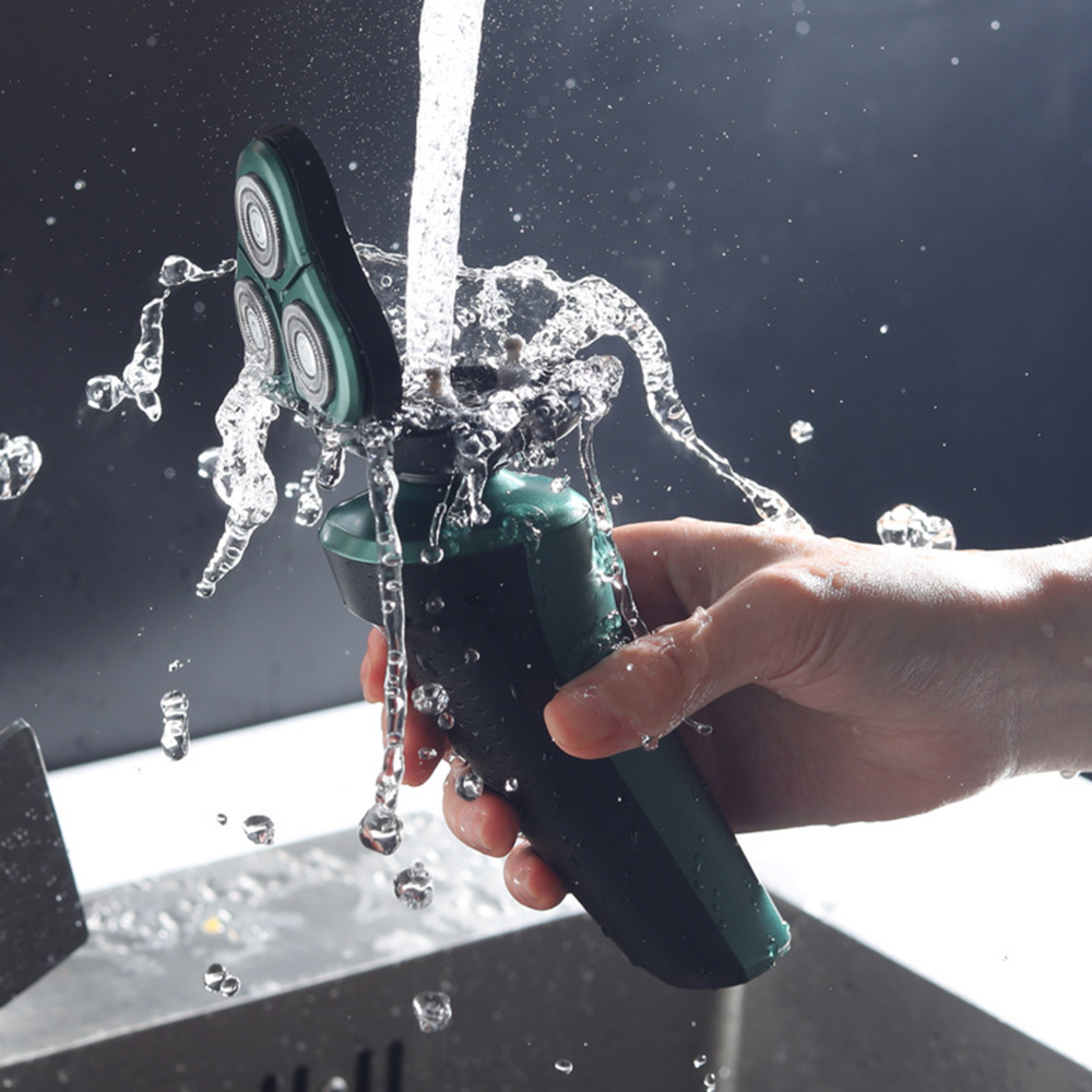 Eine Hand hält einen grünen Elektrorasierer unter fließendes Wasser, das herumspritzt.