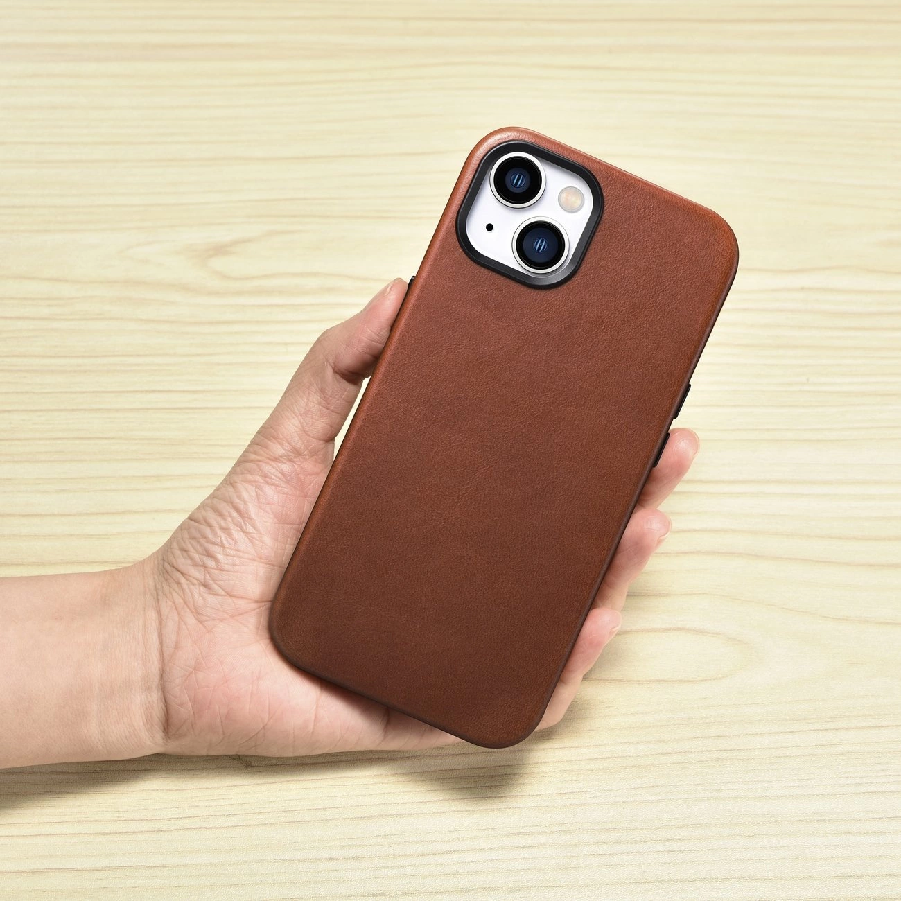 Una mano tiene uno smartphone con custodia in pelle marrone su sfondo in legno chiaro.