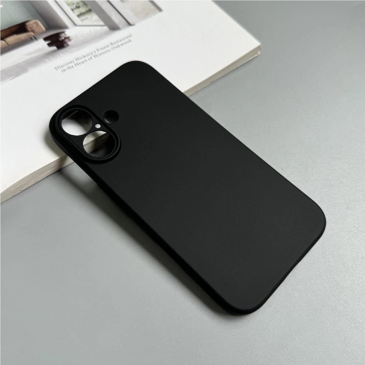 Una elegante custodia per telefono nera poggia su una superficie grigia accanto a una rivista aperta.