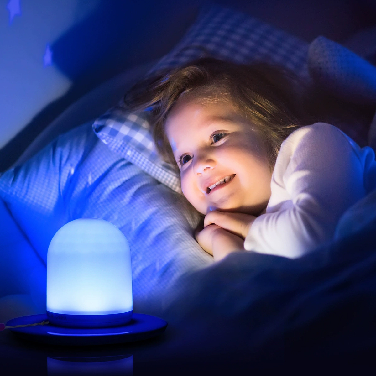 Ein kleines Kind liegt im Bett, lächelt und blickt in einer dunklen Nacht auf ein blau leuchtendes Nachtlicht.