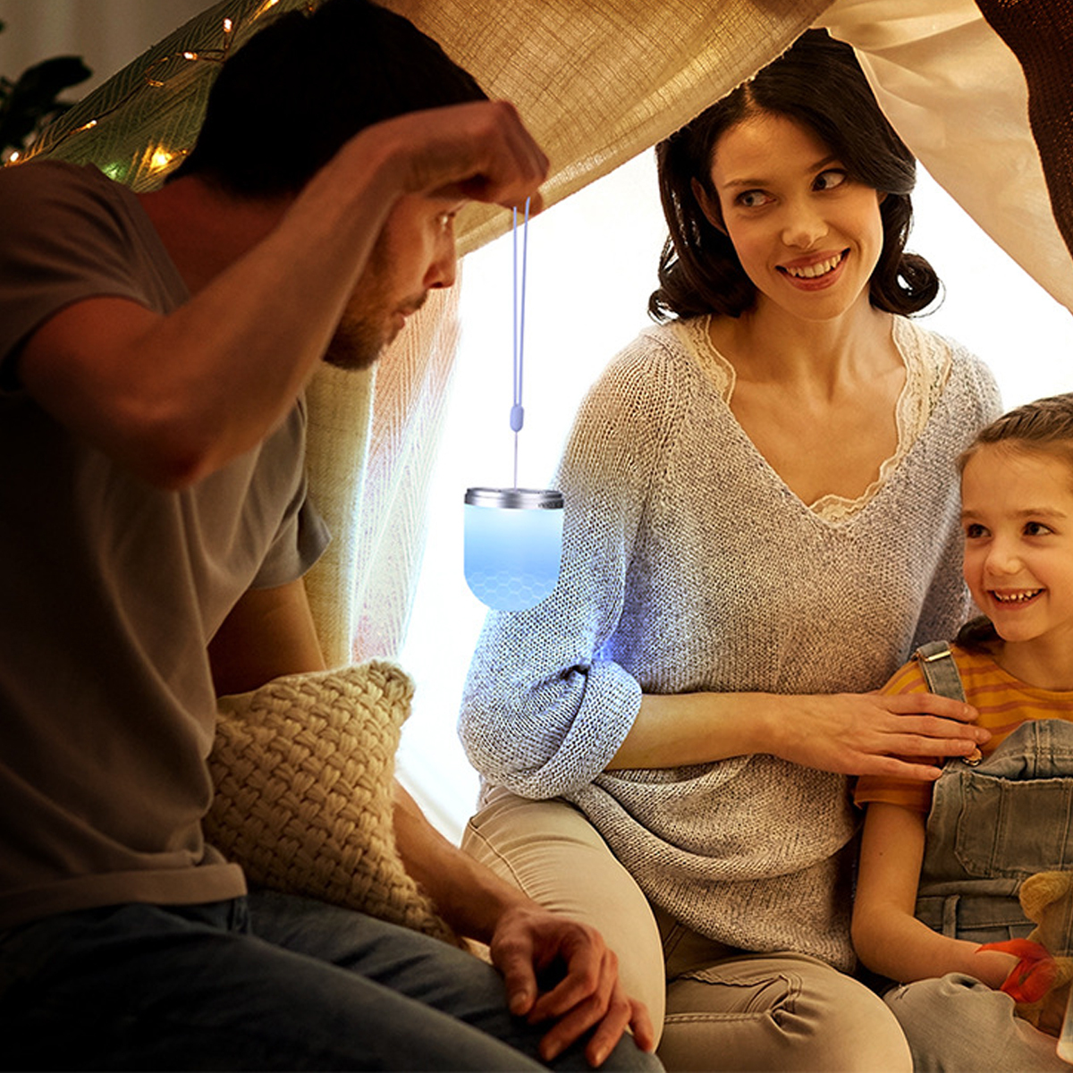 Eine Familie in einer Deckenburg, Vater hält eine blau leuchtende Hängelampe, während Mutter und Tochter lächeln.