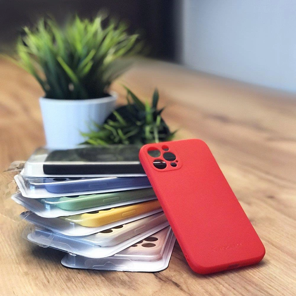 Una custodia per telefono rossa in silicone giace accanto a una pila di custodie colorate su un tavolo di legno, con una pianta.