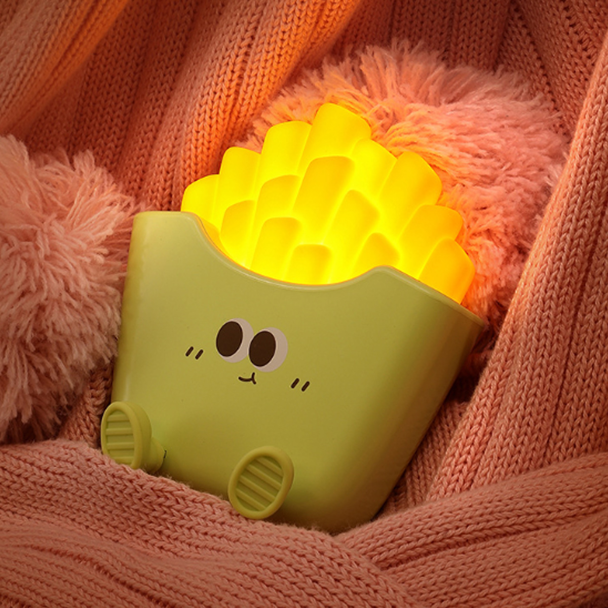 Eine niedliche, leuchtende Nachtlampe im Pommes-Frites-Design sitzt zwischen weichen, rosa Strickdecken.