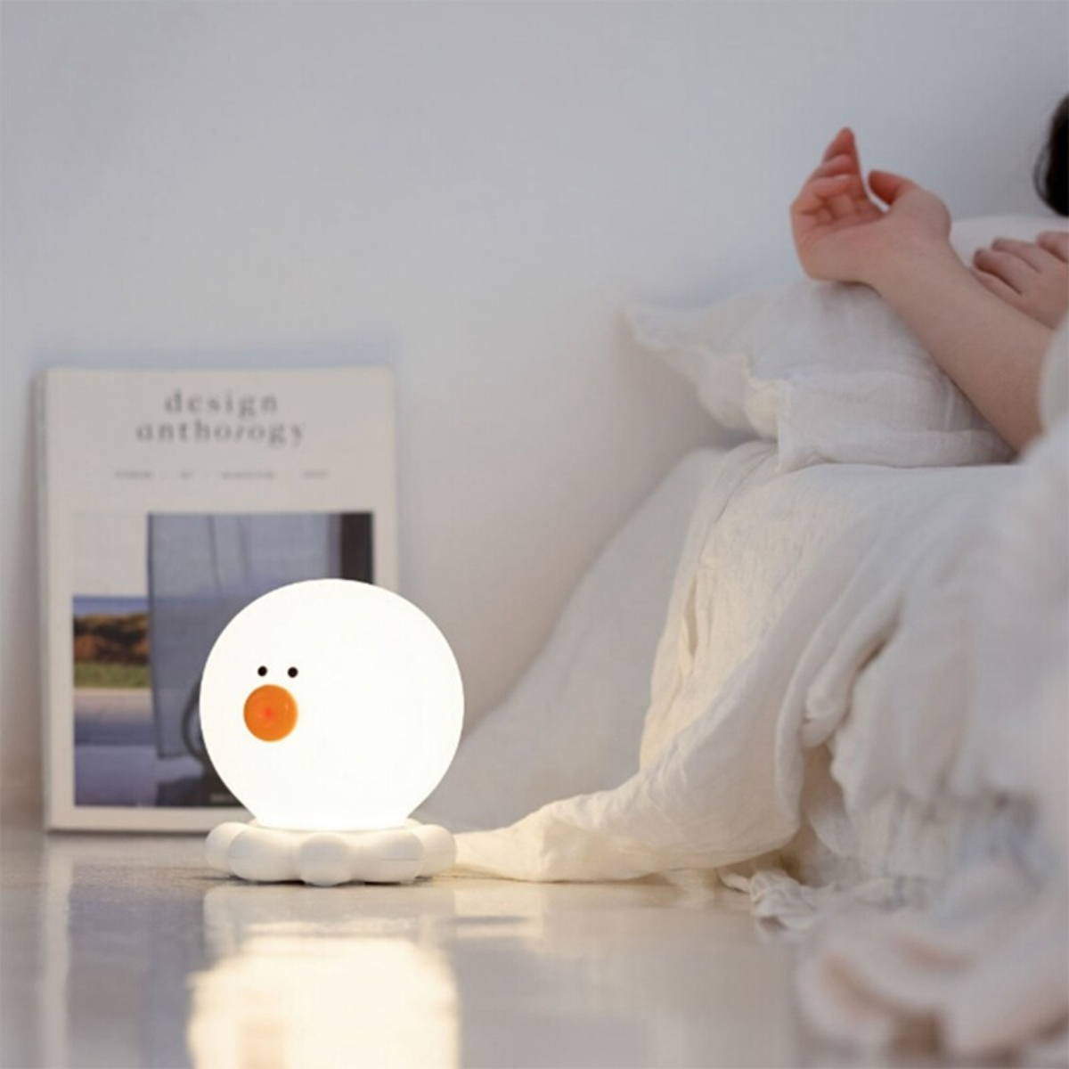 Eine weiße, runde Entenlampe leuchtet auf dem Boden neben einem Bett mit weißer Bettwäsche und einem Buch im Hintergrund.