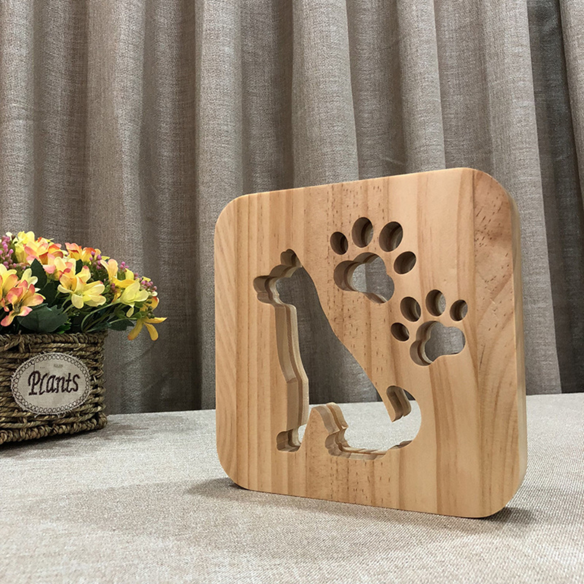 Holzblock mit ausgeschnittener Katzenform und Pfotenabdruck vor Vorhängen und Kunstblumen.