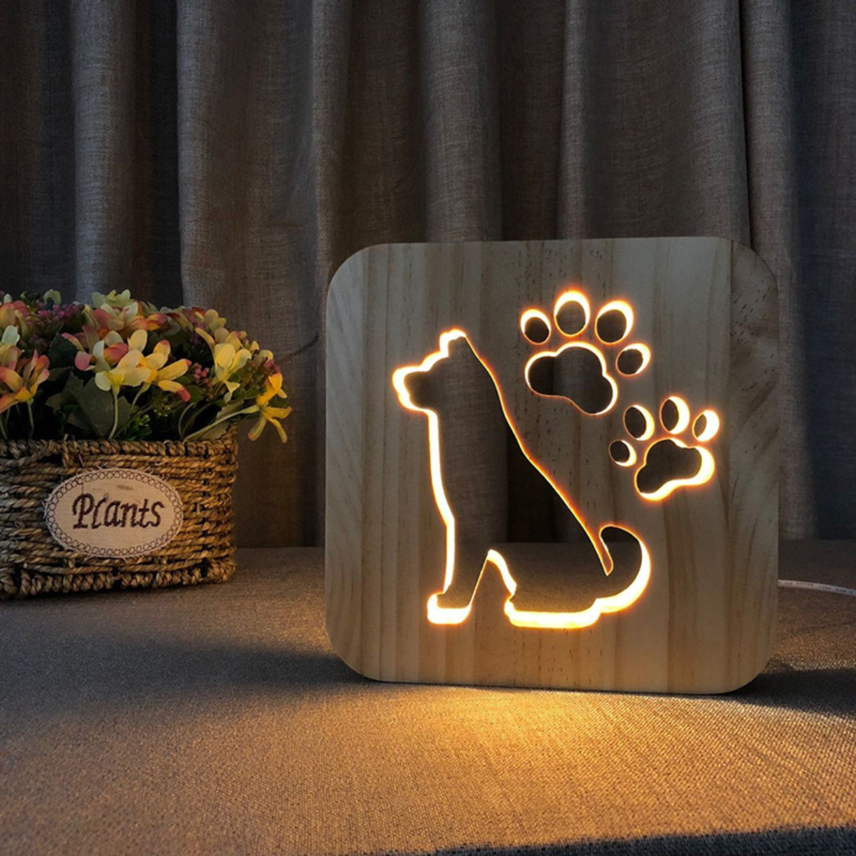 Eine beleuchtete Holzlampe mit Hunde- und Pfotenabdruck-Ausschnitt steht vor einem Vorhang neben Blumen.