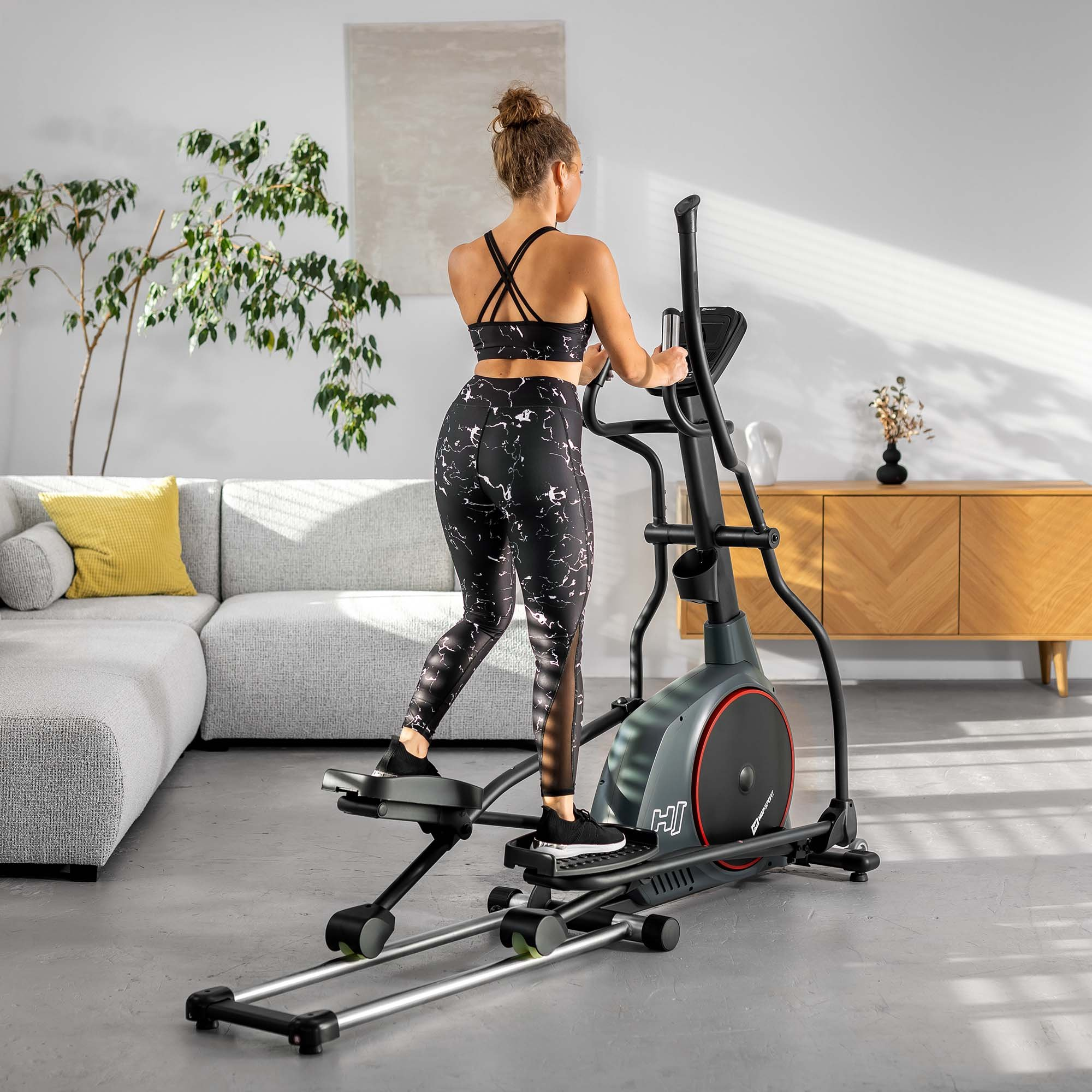 Eine Frau in Sportkleidung trainiert zu Hause auf einem schwarzen Crosstrainer in einem hellen Wohnzimmer.