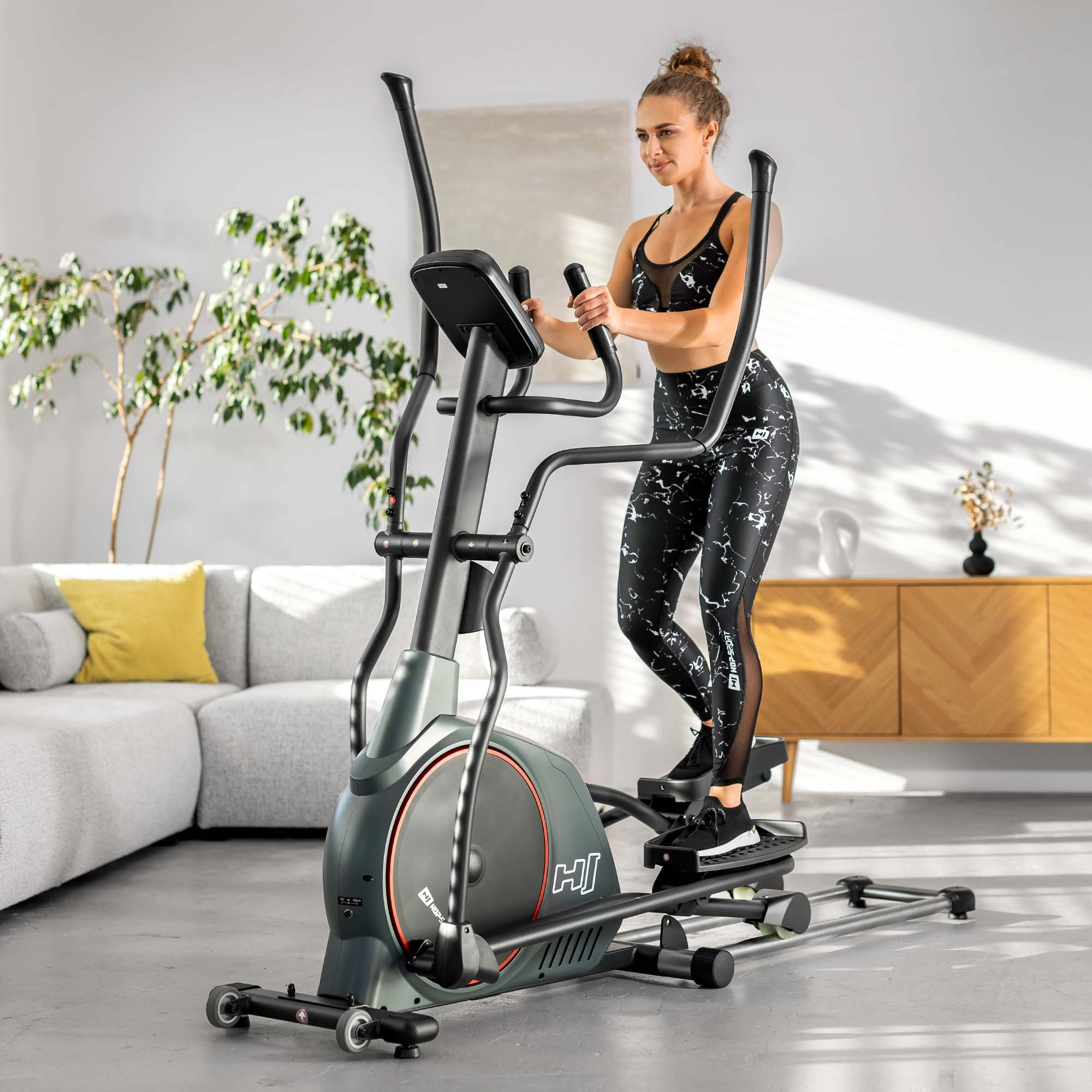 Eine Frau in einem passenden schwarzen Trainingsset trainiert auf einem grauen und schwarzen Crosstrainer im Wohnzimmer.