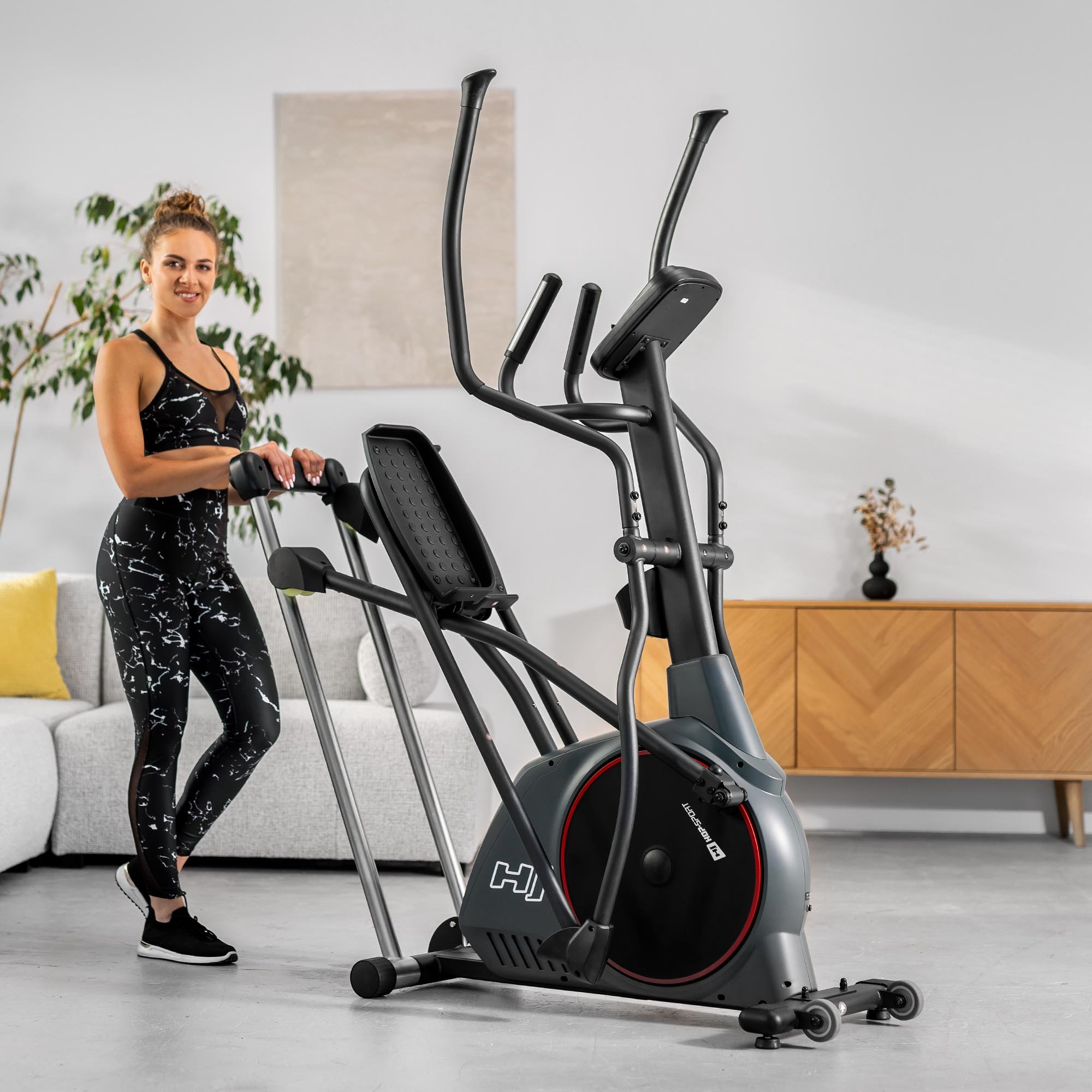 Eine Frau in Sportkleidung lächelt und lehnt sich an einen Crosstrainer in einem hellen Wohnzimmer.