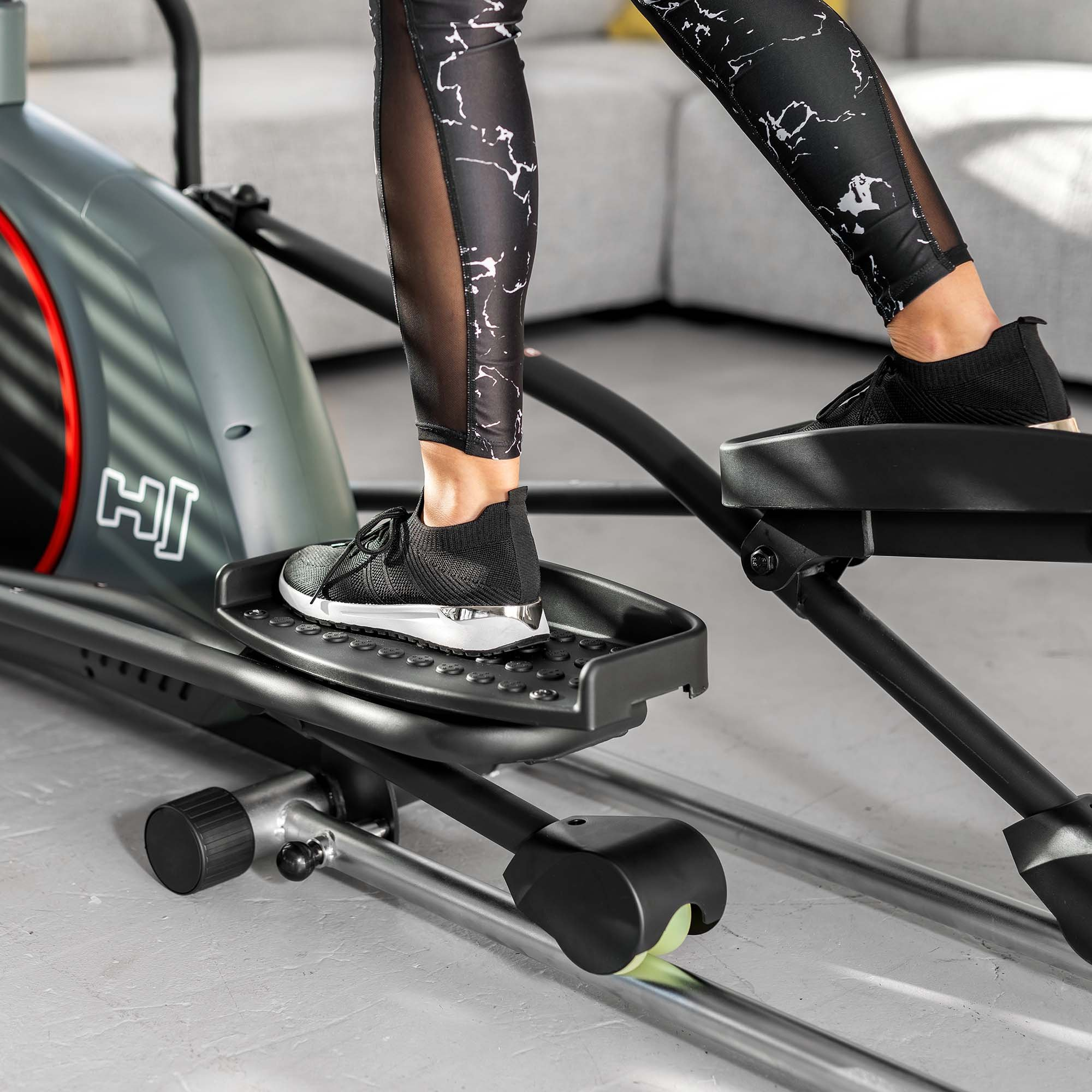 Person in gemusterten Leggings trainiert auf einem grauen Crosstrainer in einem Raum.
