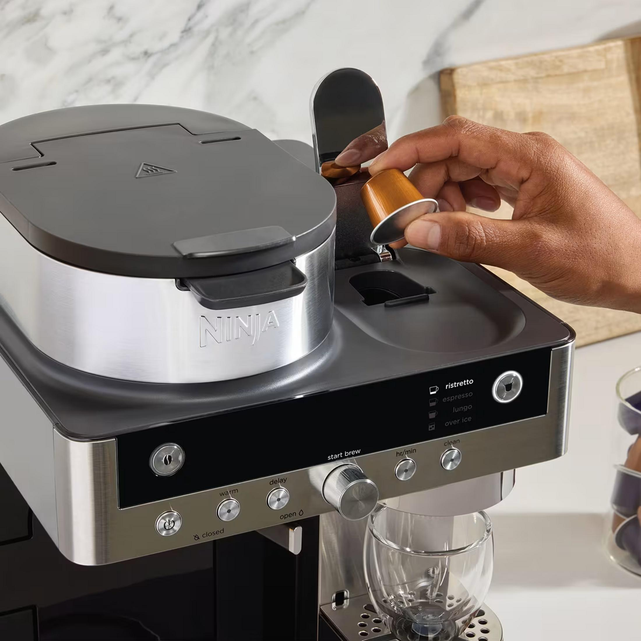 Una mano inserisce una capsula di caffè dorata in una moderna macchina da caffè Ninja su un bancone da cucina.