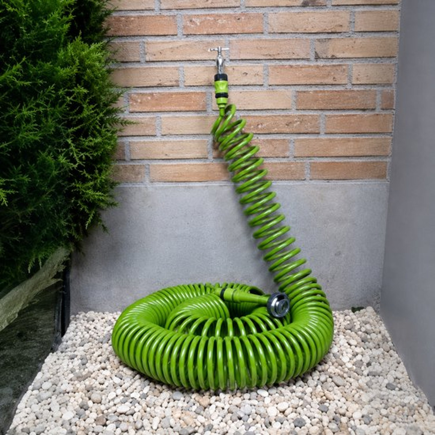 A bright green spiral garden hose is connected to a spigot on a brick wall, resting on white pebbles.