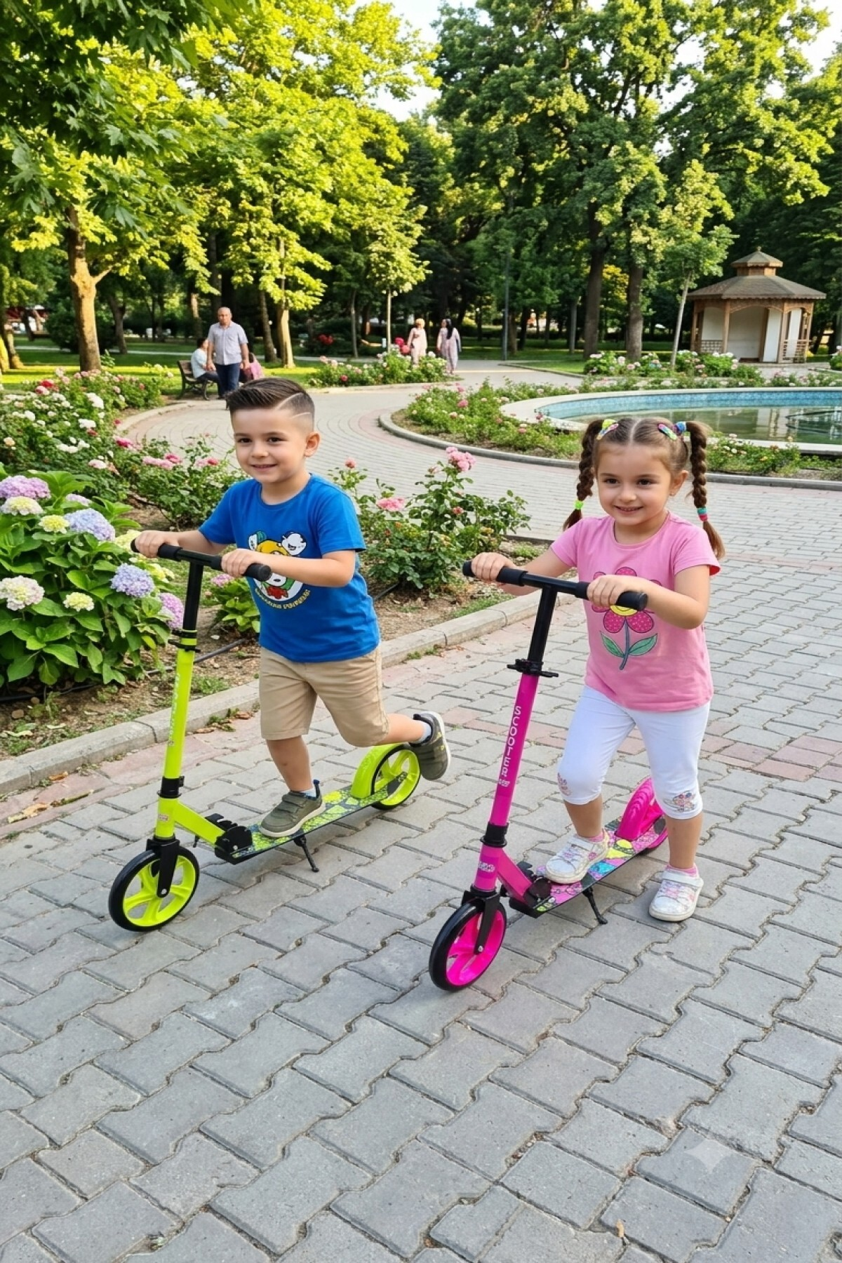 Küçük bir çocuk ve kız, ağaçlar ve çiçeklerle çevrili park yolunda renkli scooter'lara biniyor.