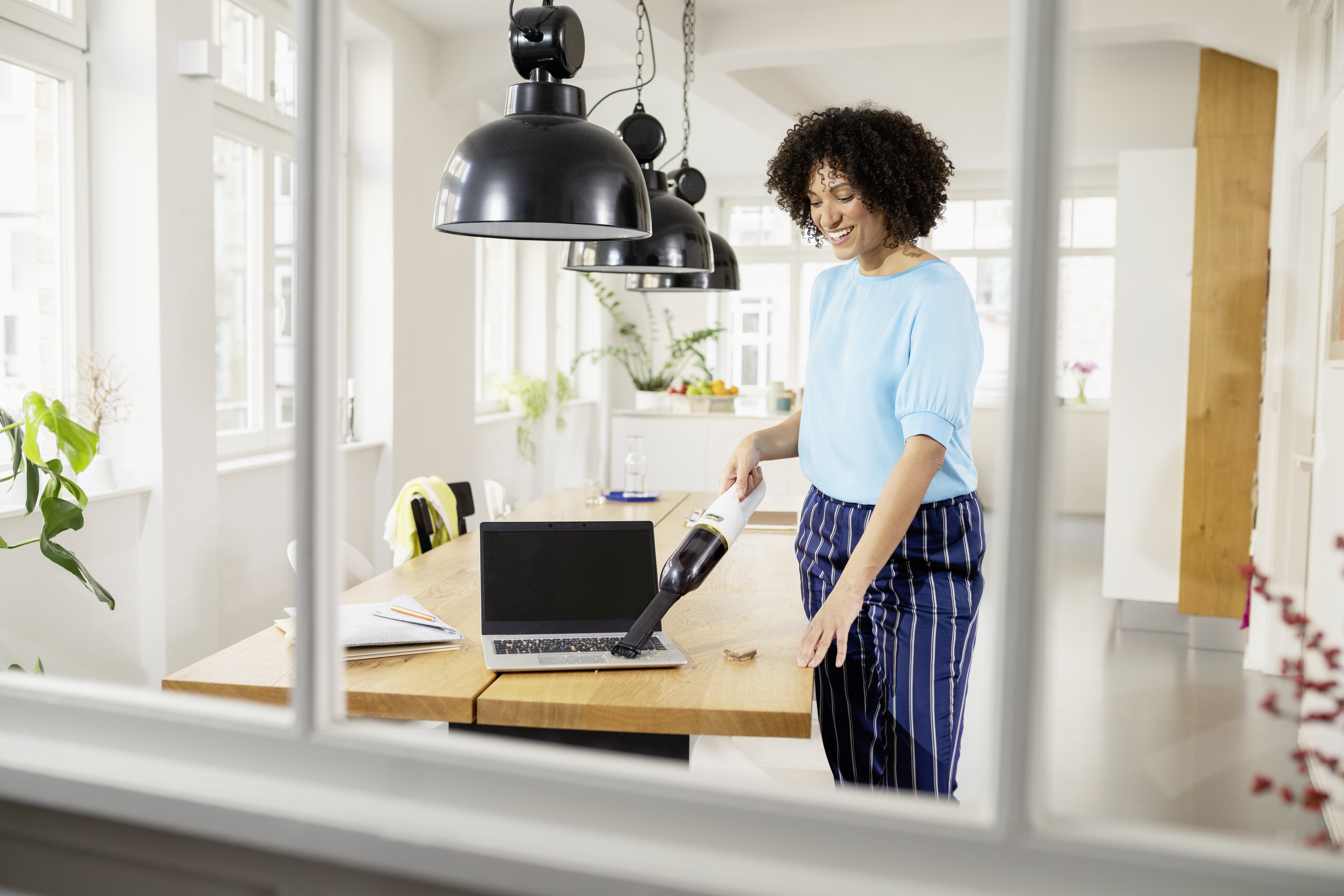Een vrouw in een blauwe top zuigt kruimels op van een laptop op een houten tafel.