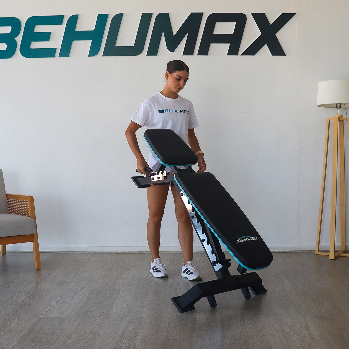 Una mujer con camiseta blanca ajusta un banco de pesas ajustable frente a una pared con el logo Behumax.