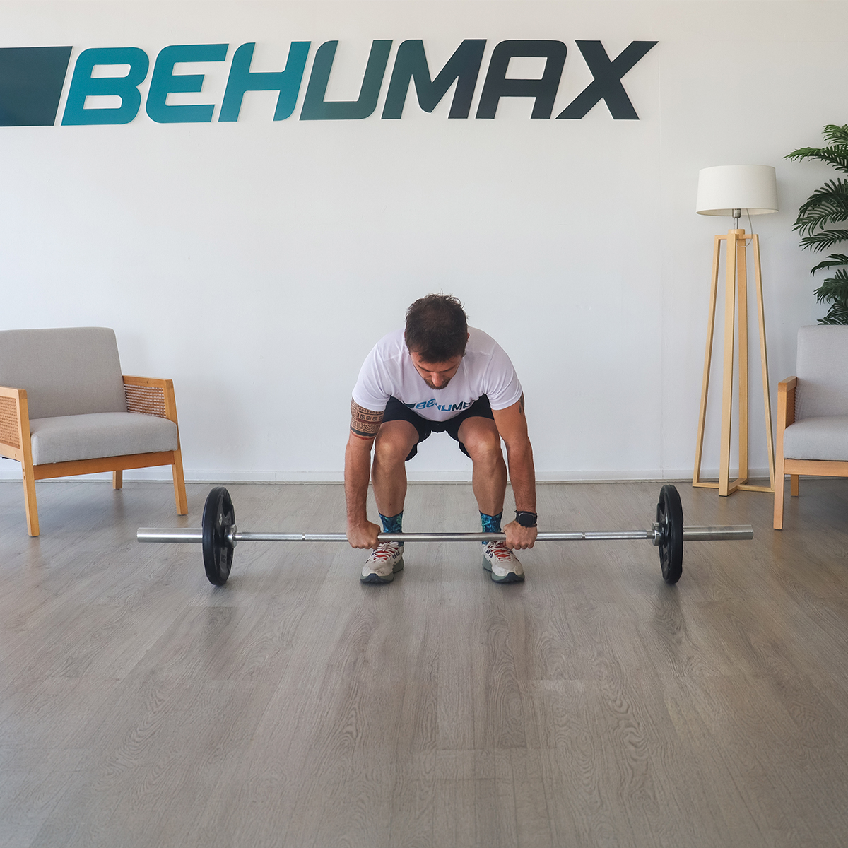 Un hombre con ropa deportiva se prepara para levantar una barra en una sala con un logo BEHUMAX.