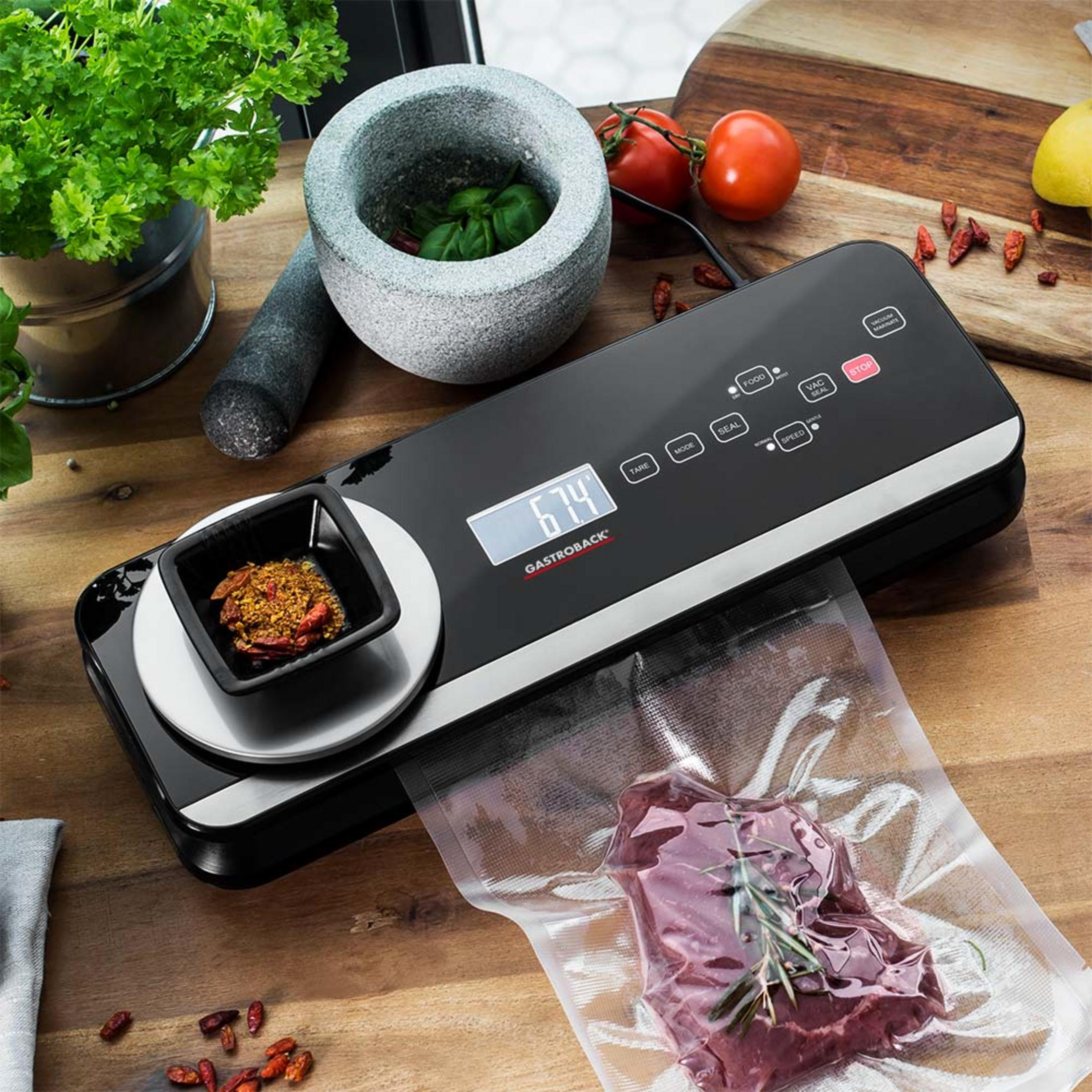 Una macchina sottovuoto nera Gastroback con bilancia su un tavolo di legno, vicino a carne, erbe e pomodori.