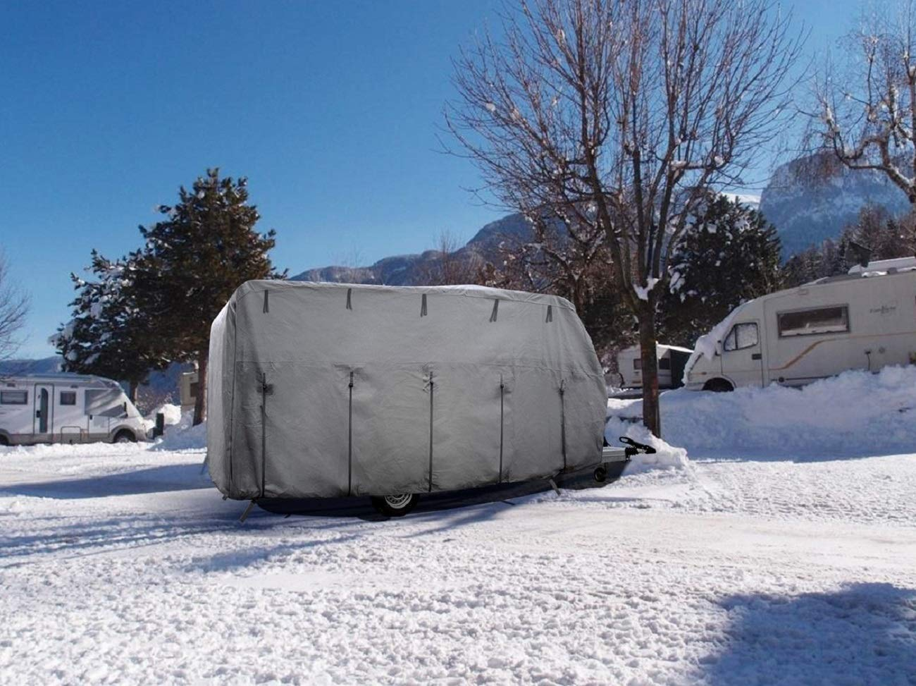 Ein grauer Wohnwagen mit Schutzhülle steht im Schnee vor Bergen.