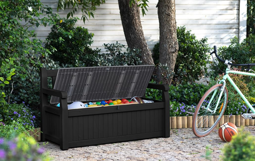 Eine schwarze Aufbewahrungsbank mit offenem Deckel steht auf einer Terrasse im Garten, ein hellgrünes Fahrrad daneben.