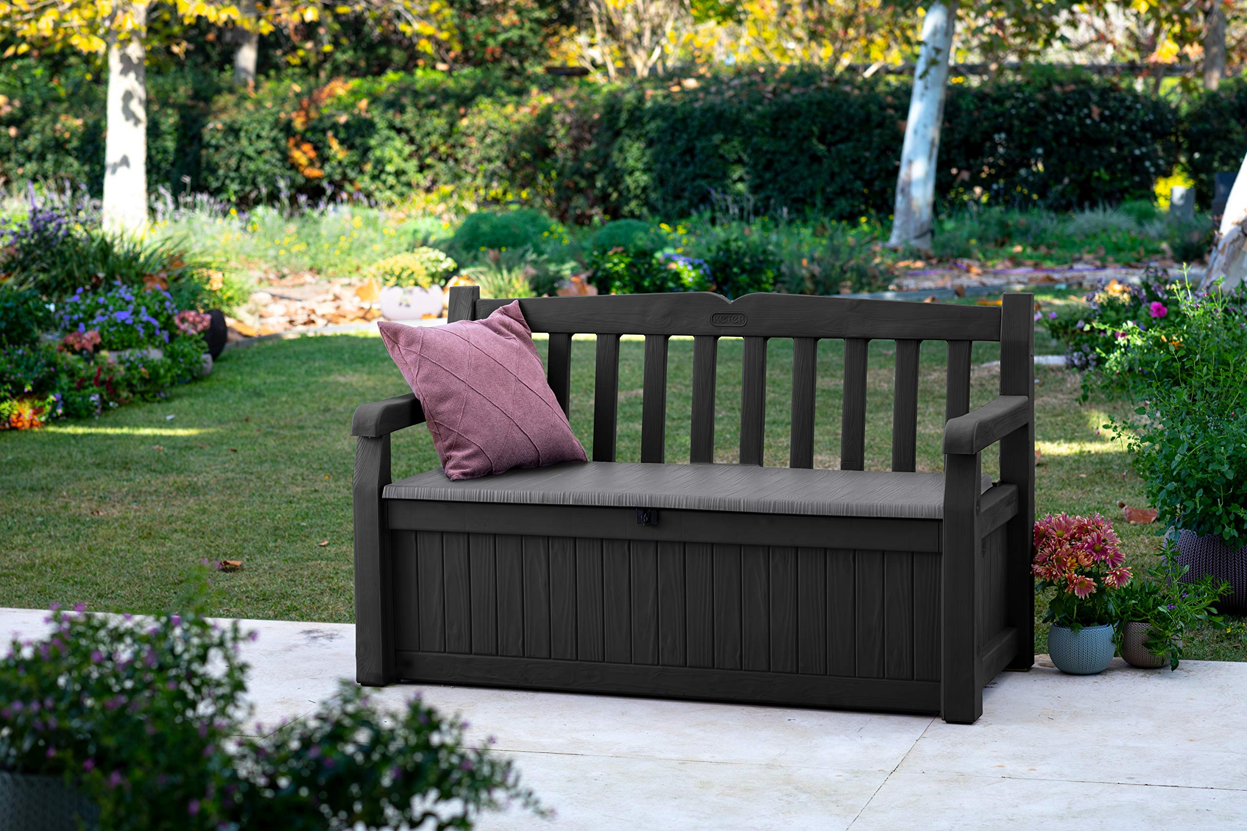 Eine dunkelgraue Gartenbank mit Stauraum und Kissen steht auf einer Terrasse vor einer grünen Wiese und Blumen.