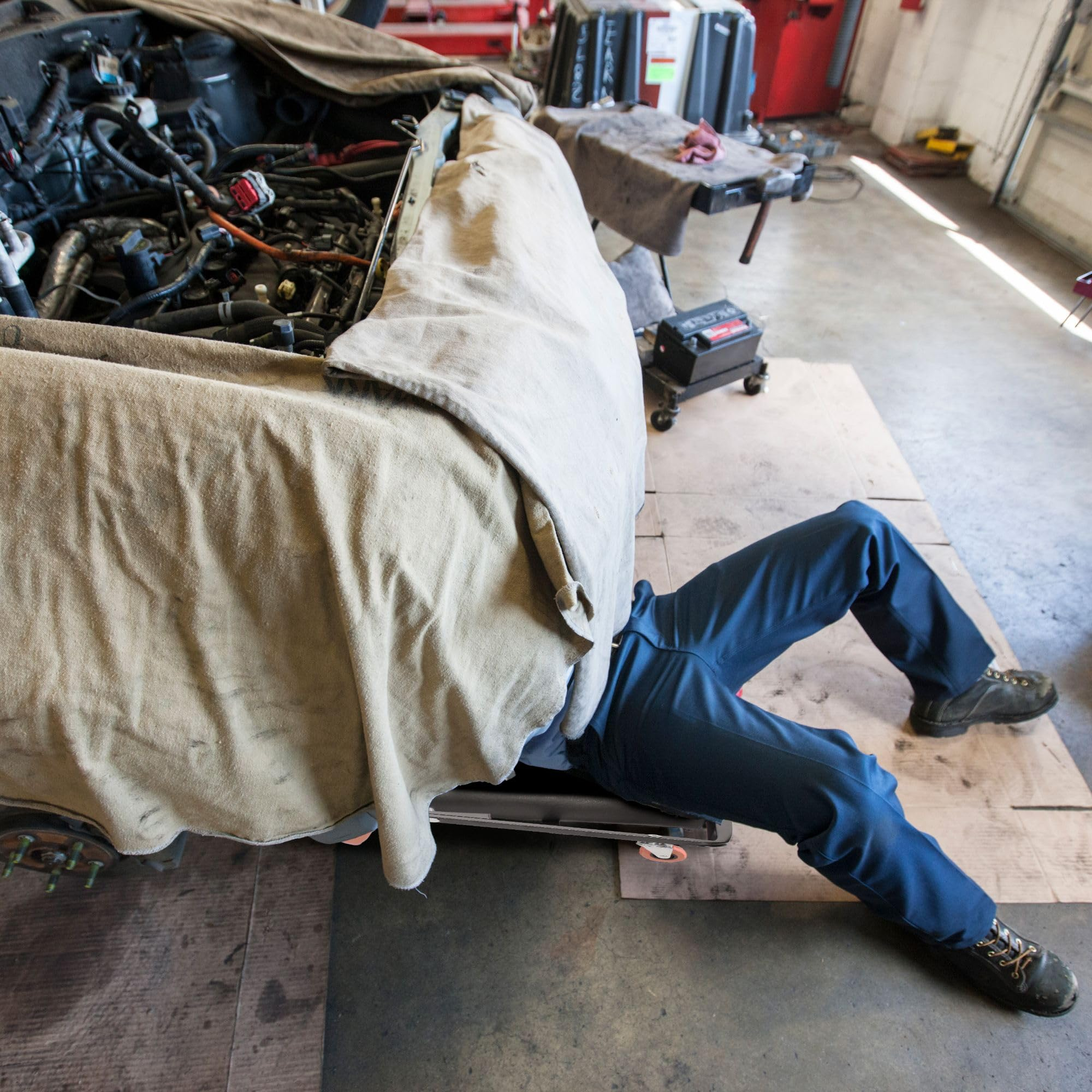 Mechaniker in blauer Hose arbeitet unter einem mit Plane bedeckten Auto.