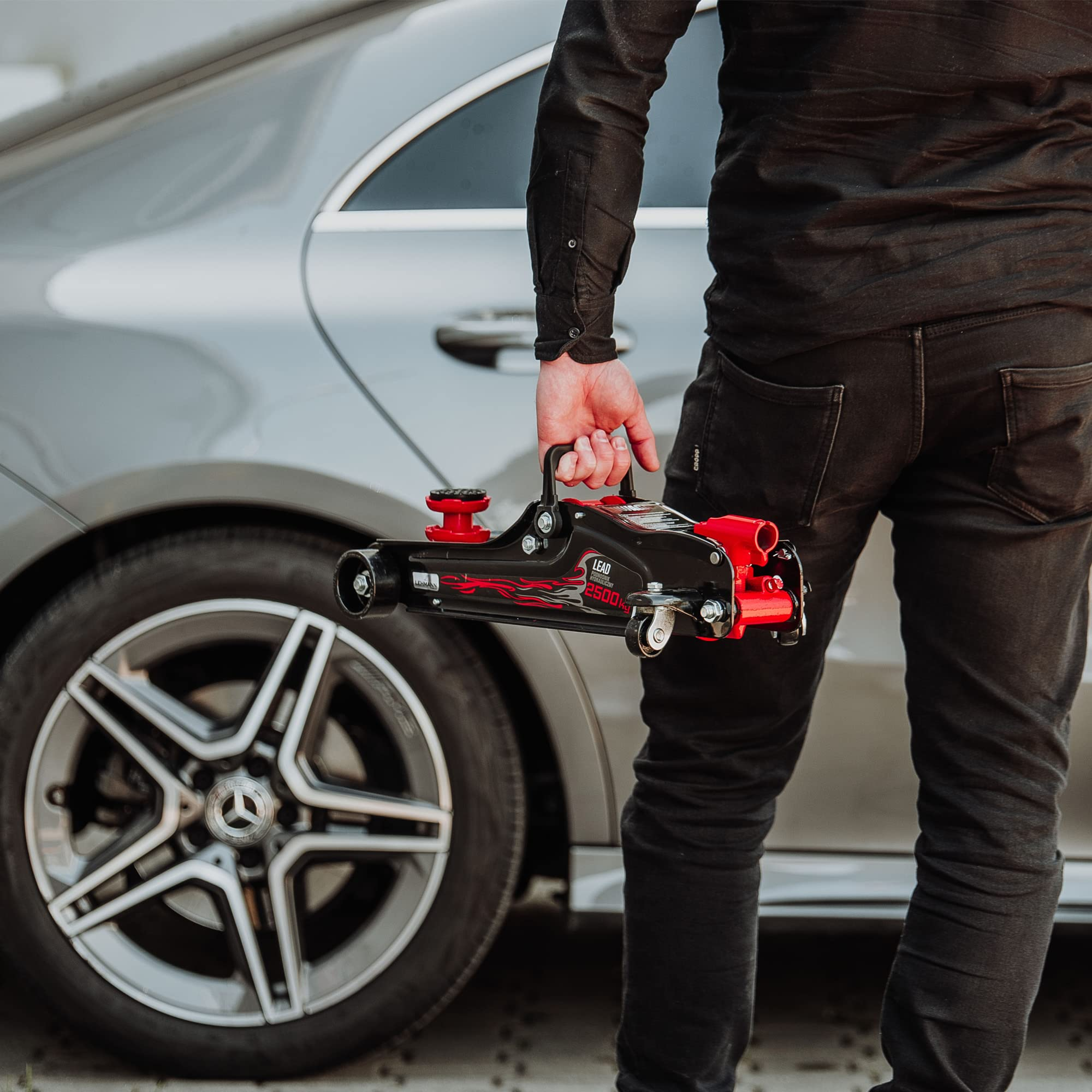 Un uomo in nero tiene un cric per auto rosso e nero davanti a un'auto.