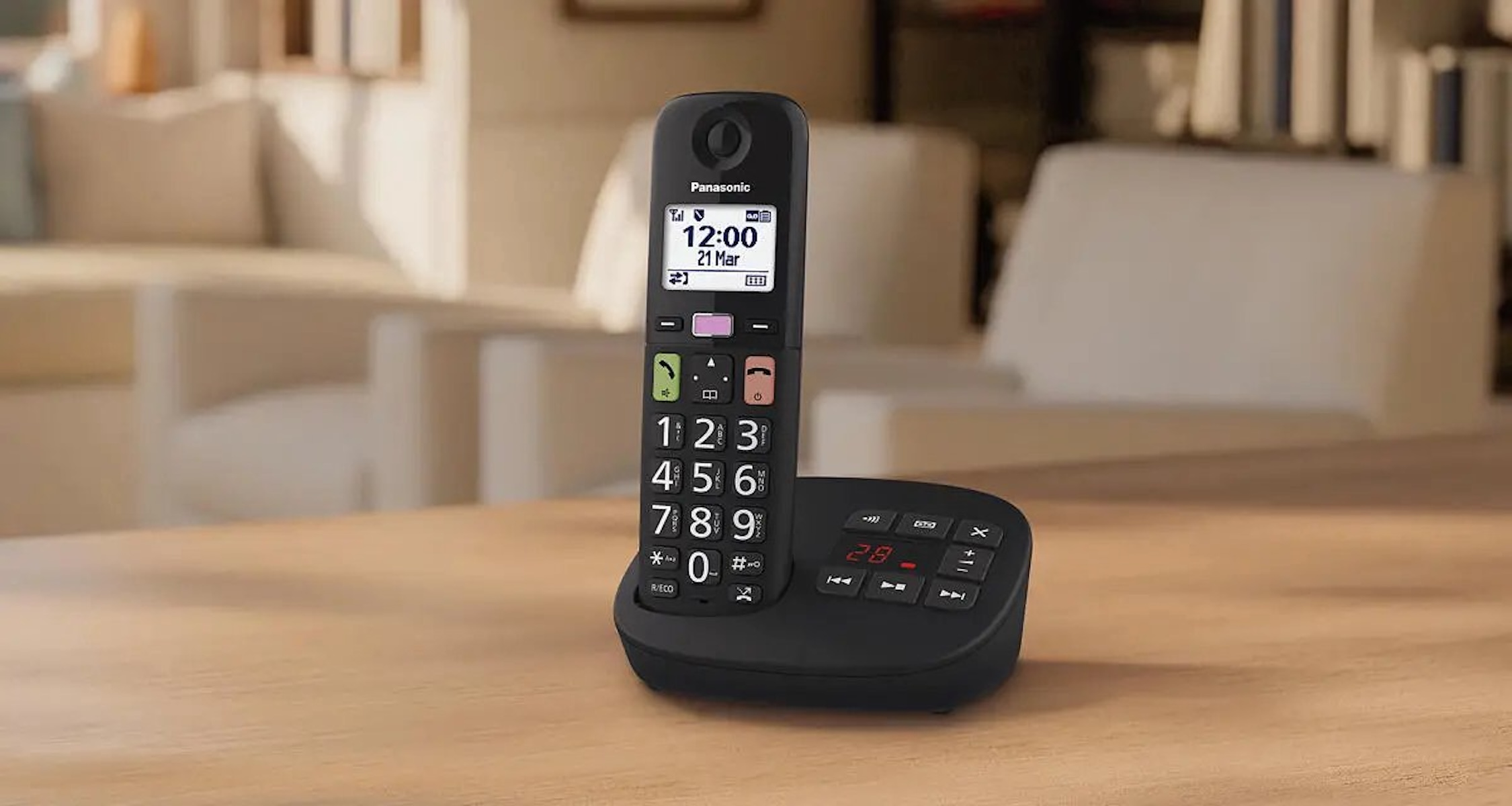 Un téléphone fixe sans fil Panasonic noir sur sa base de charge, posé sur une table en bois dans un salon lumineux.