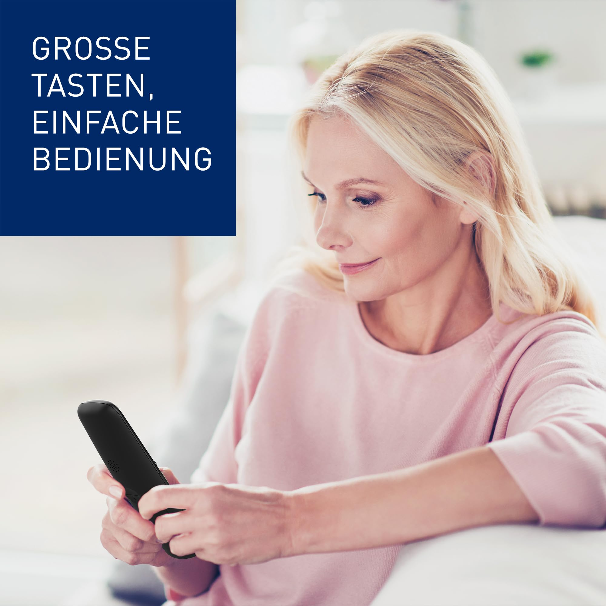Une femme blonde est assise sur un canapé et tient un téléphone portable noir.