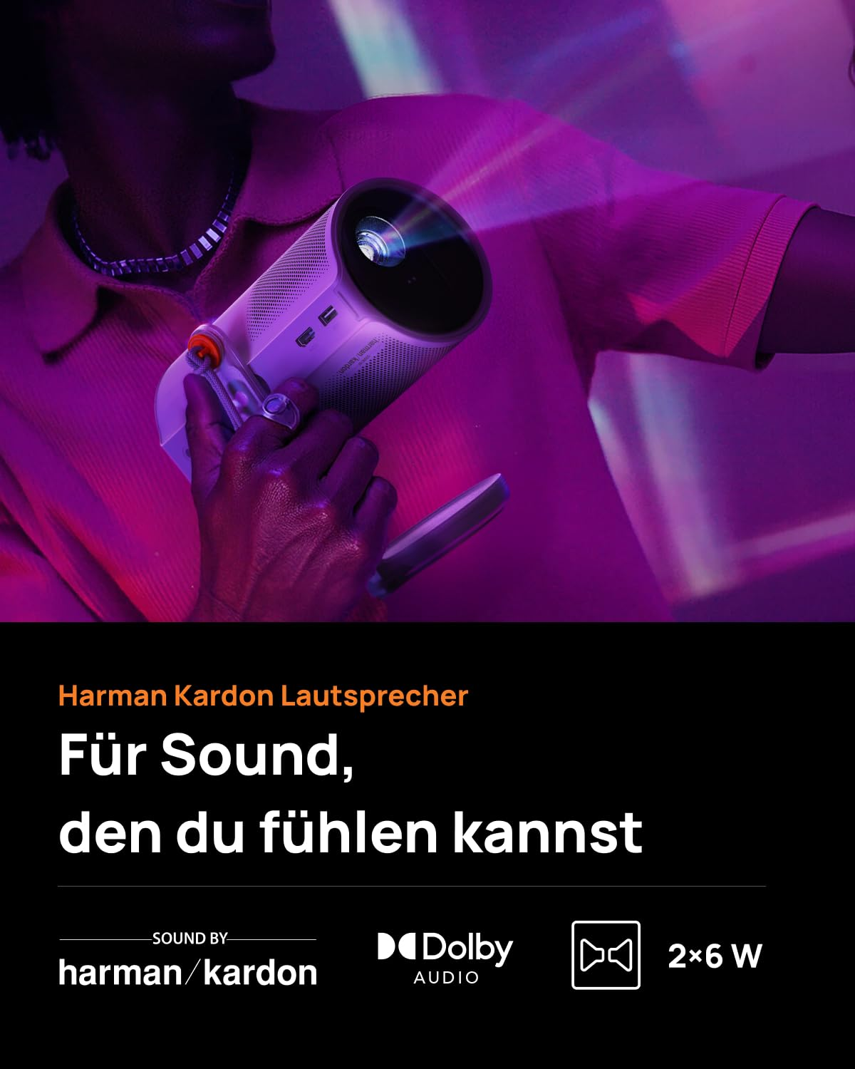 Eine Person hält einen kleinen weißen Projektor mit Harman Kardon Lautsprecher vor einem violetten Hintergrund.