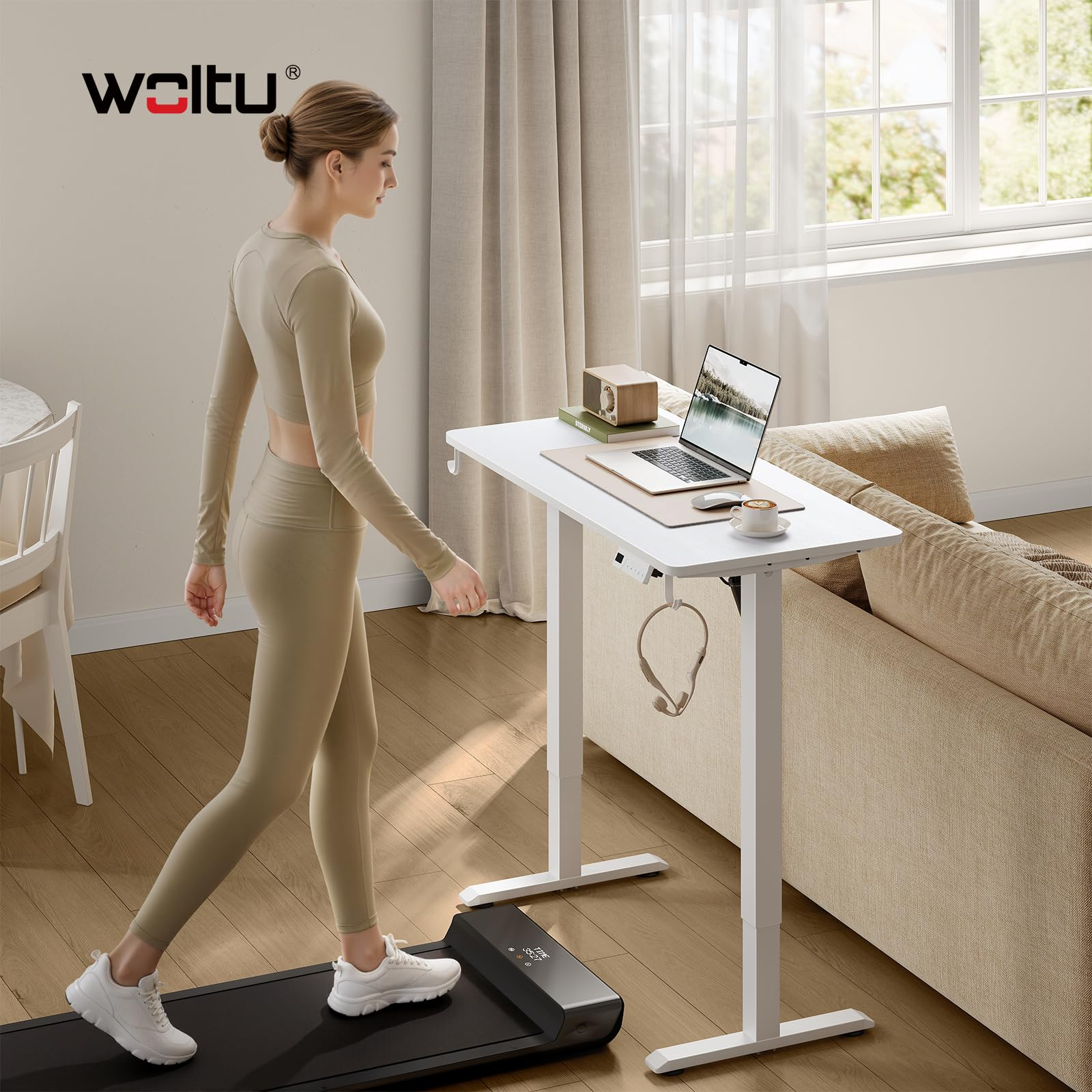 Une femme marche sur un tapis de course près d'un bureau réglable blanc avec un ordinateur portable et un canapé.