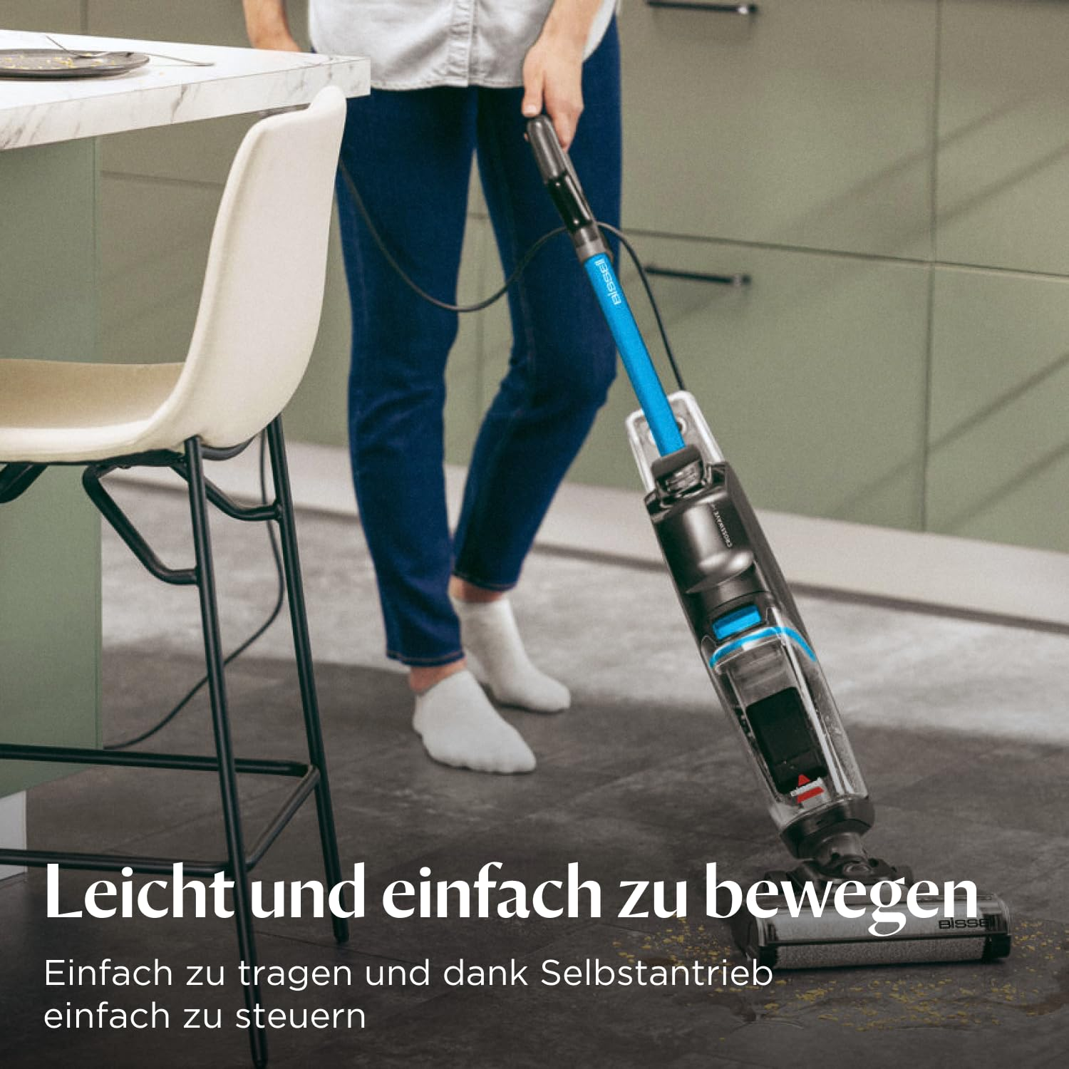 Persona in jeans e calzini bianchi pulisce il pavimento della cucina con un pulitore blu e nero.