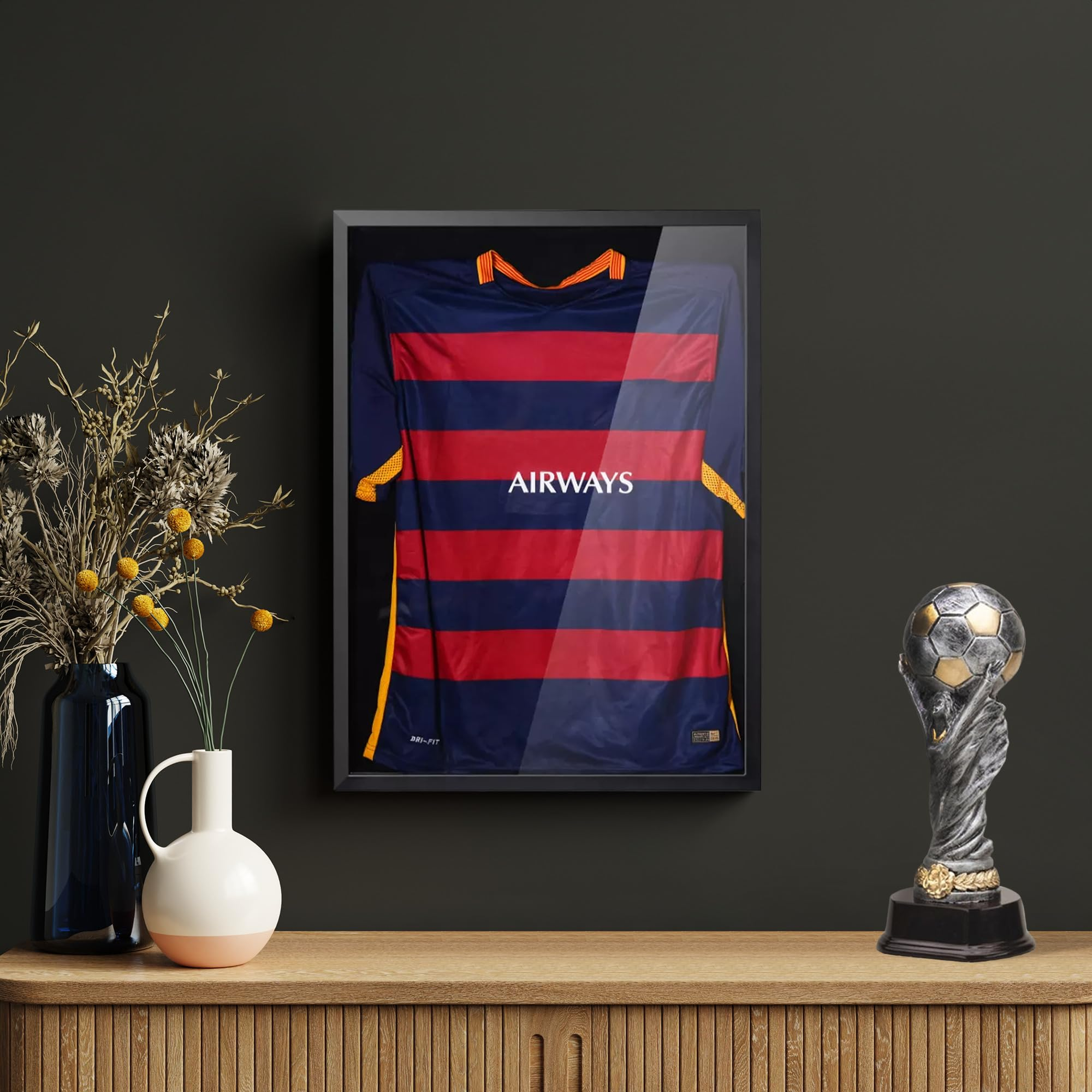 Ein gerahmtes Trikot, eine Vase mit Blumen und ein Pokal stehen auf einem Holzkabinett vor einer dunklen Wand.