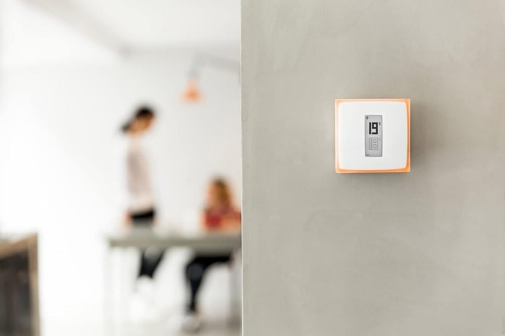 Ein weißer, quadratischer Thermostat mit orangem Rand an einer hellgrauen Wand, im Hintergrund zwei verschwommene Personen.
